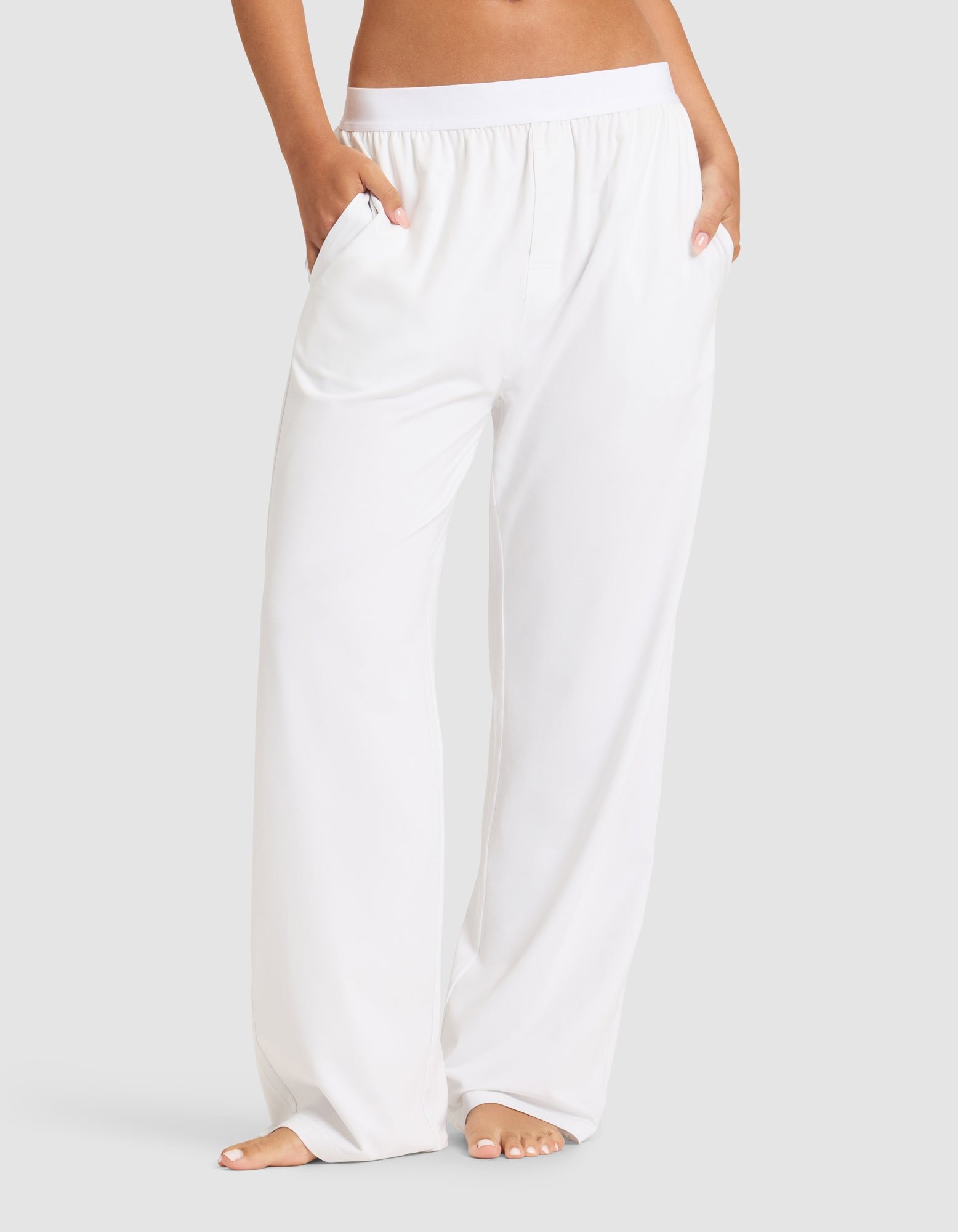 A woman stands barefoot, hands in pockets, wearing Cozy Earth's Women's First Layer Pajama Pant—loose-fitting white pants with an elastic waistband—against a plain light gray background.