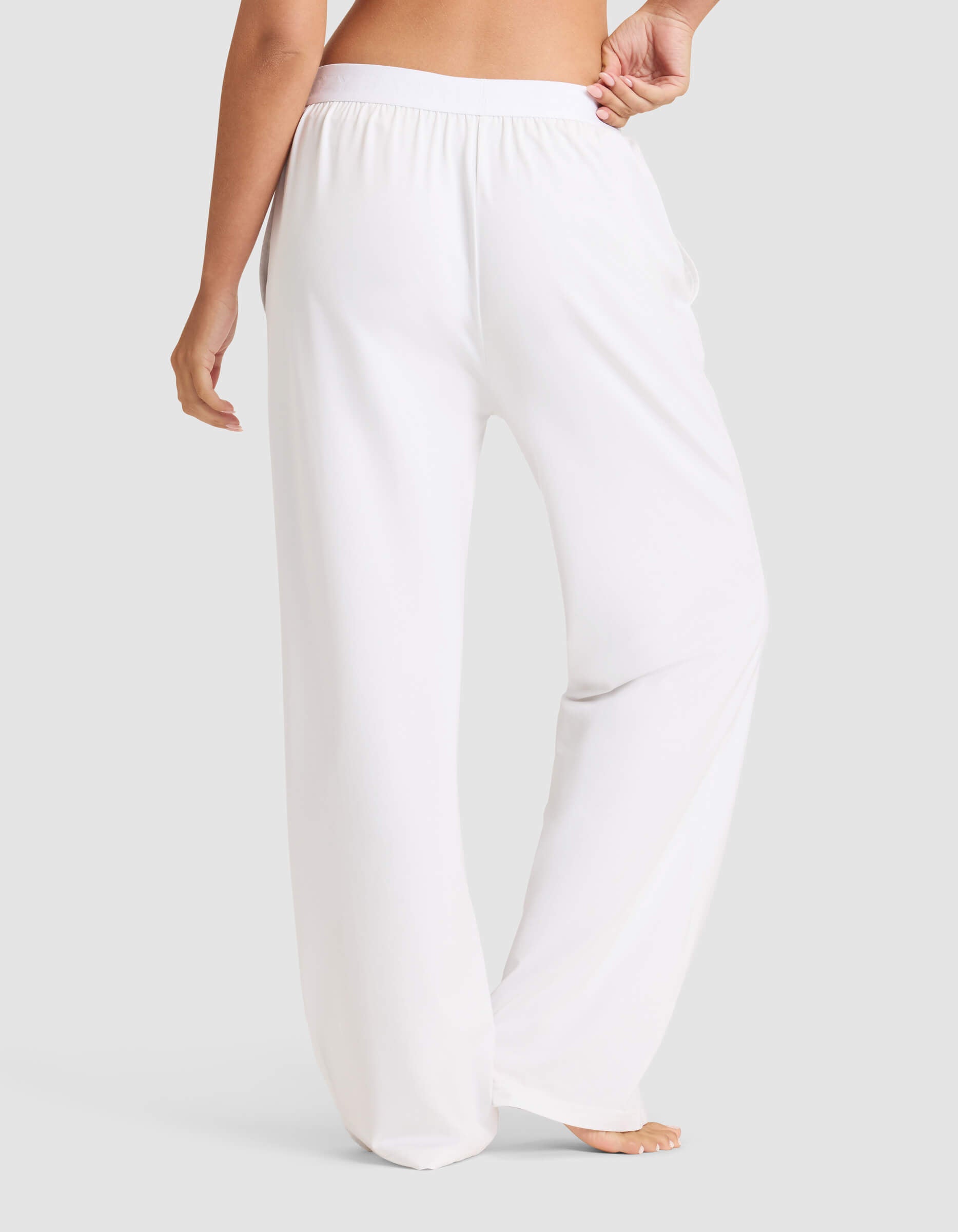 A woman stands barefoot, shown from the back against a plain light gray background, wearing Cozy Earth's Women's First Layer Pajama Pant—loose-fitting white bottoms with an elastic waistband.|Color:White