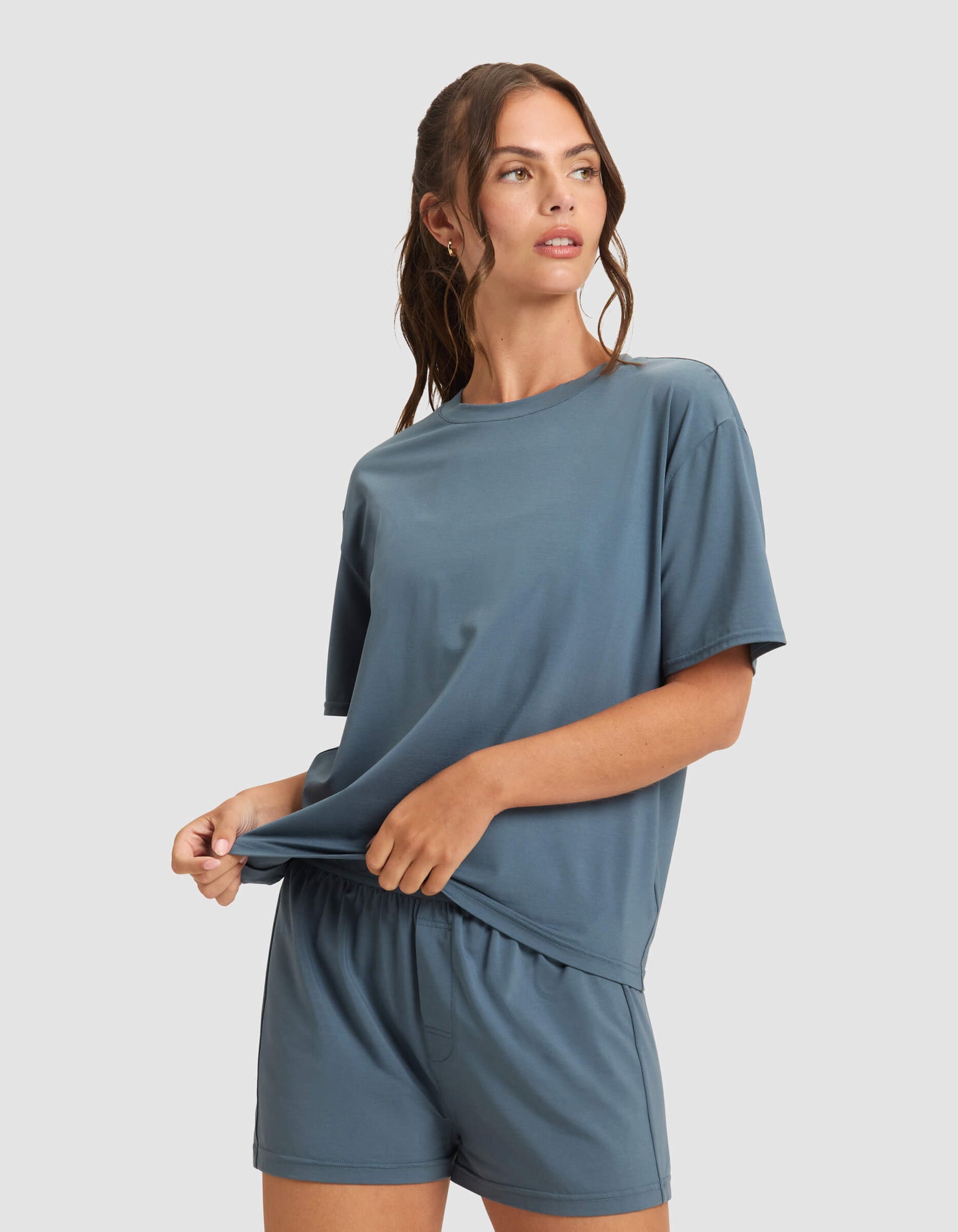 A woman models the Cozy Earth Women's First Layer Pajama Short Sleeve Tee & Boxer Short Set in blue, standing against a light gray background while holding the hem of her T-shirt and looking to the side.