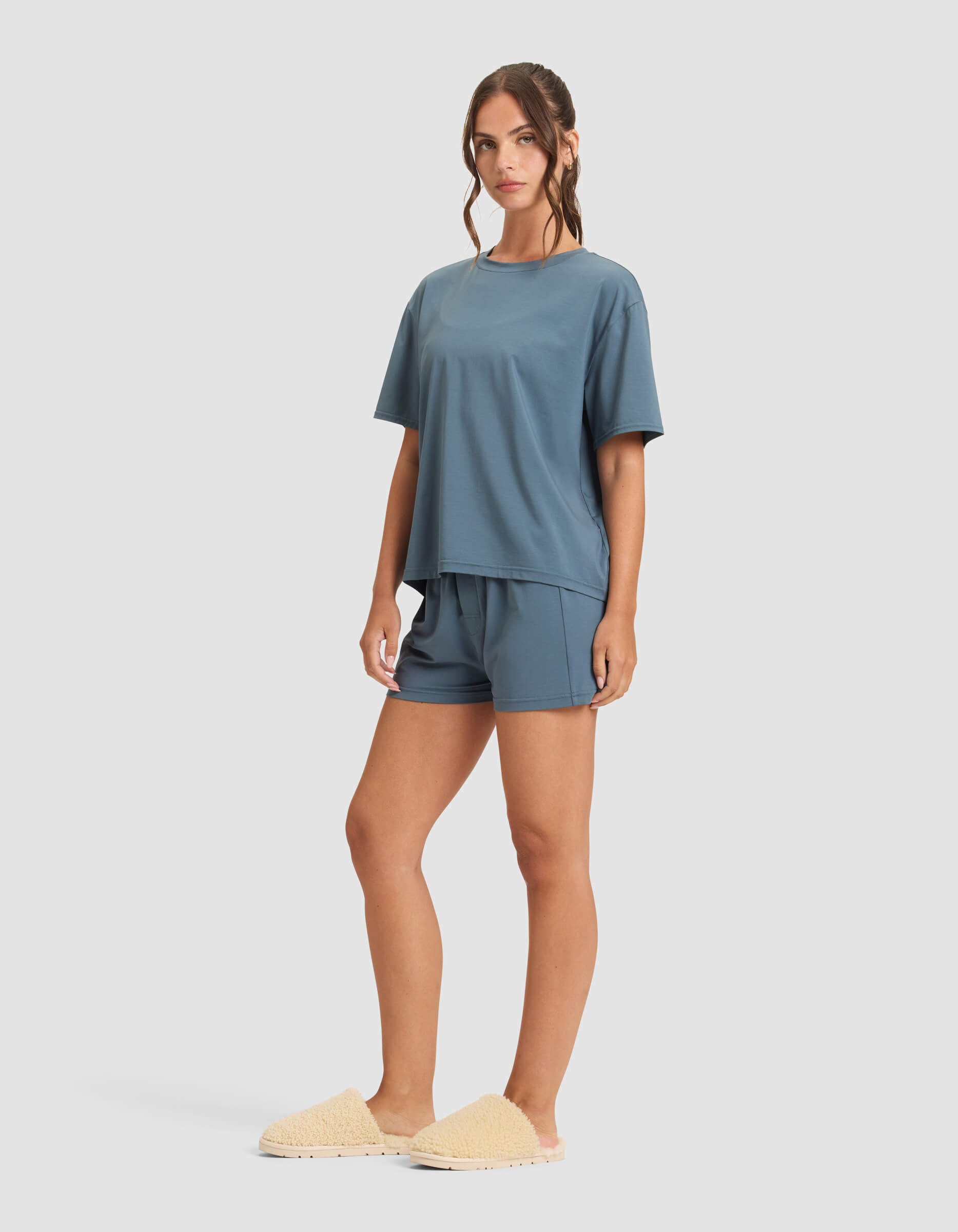 A woman models the Cozy Earth Women's First Layer Pajama Short Sleeve Tee & Boxer Short Set in blue, paired with beige fuzzy slippers, against a plain light background.|Color:Dawn