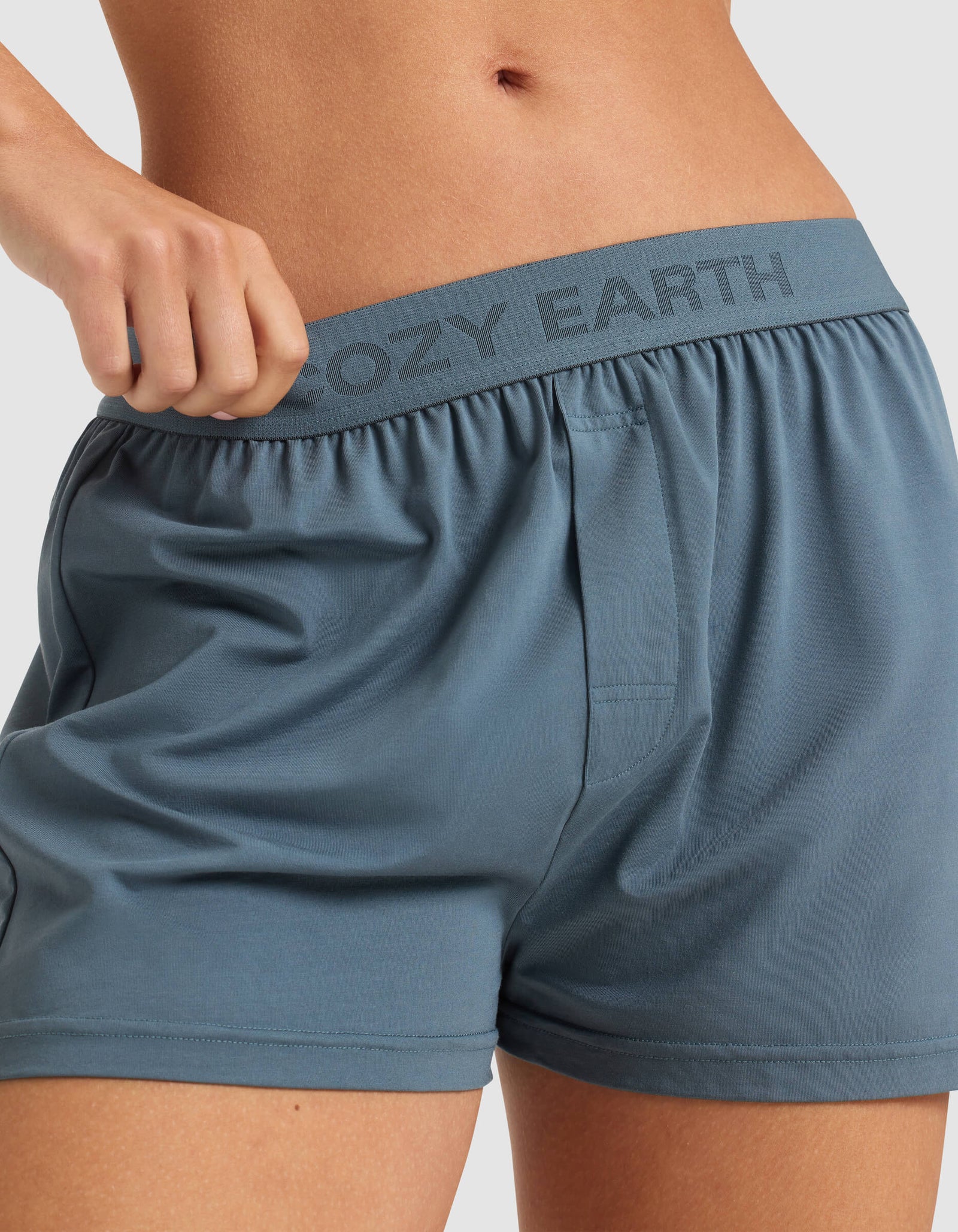 A person wears light blue Cozy Earth Women's First Layer Pajama Short Sleeve Tee & Boxer Short Set, pulling at the waistband to reveal the brand name on the elastic. The cropped image shows only the torso and upper thighs.