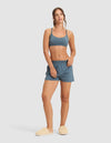 A woman poses against a plain background wearing the Cozy Earth Women's First Layer Pajama Soft Bra, matching blue shorts, and beige fuzzy slippers. She stands with one hand on her hip and gazes at the camera with a relaxed look.|Color:Dawn