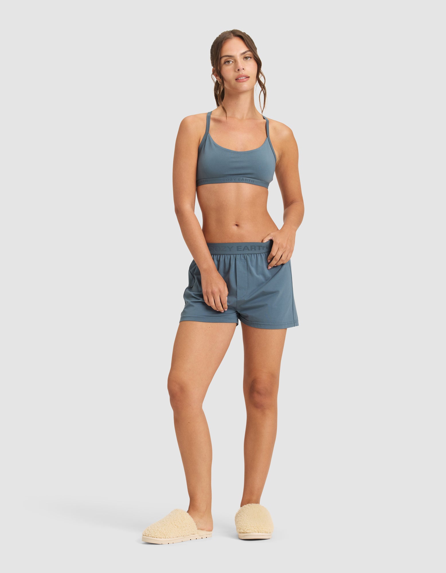 A woman poses against a plain background wearing the Cozy Earth Women's First Layer Pajama Soft Bra, matching blue shorts, and beige fuzzy slippers. She stands with one hand on her hip and gazes at the camera with a relaxed look.