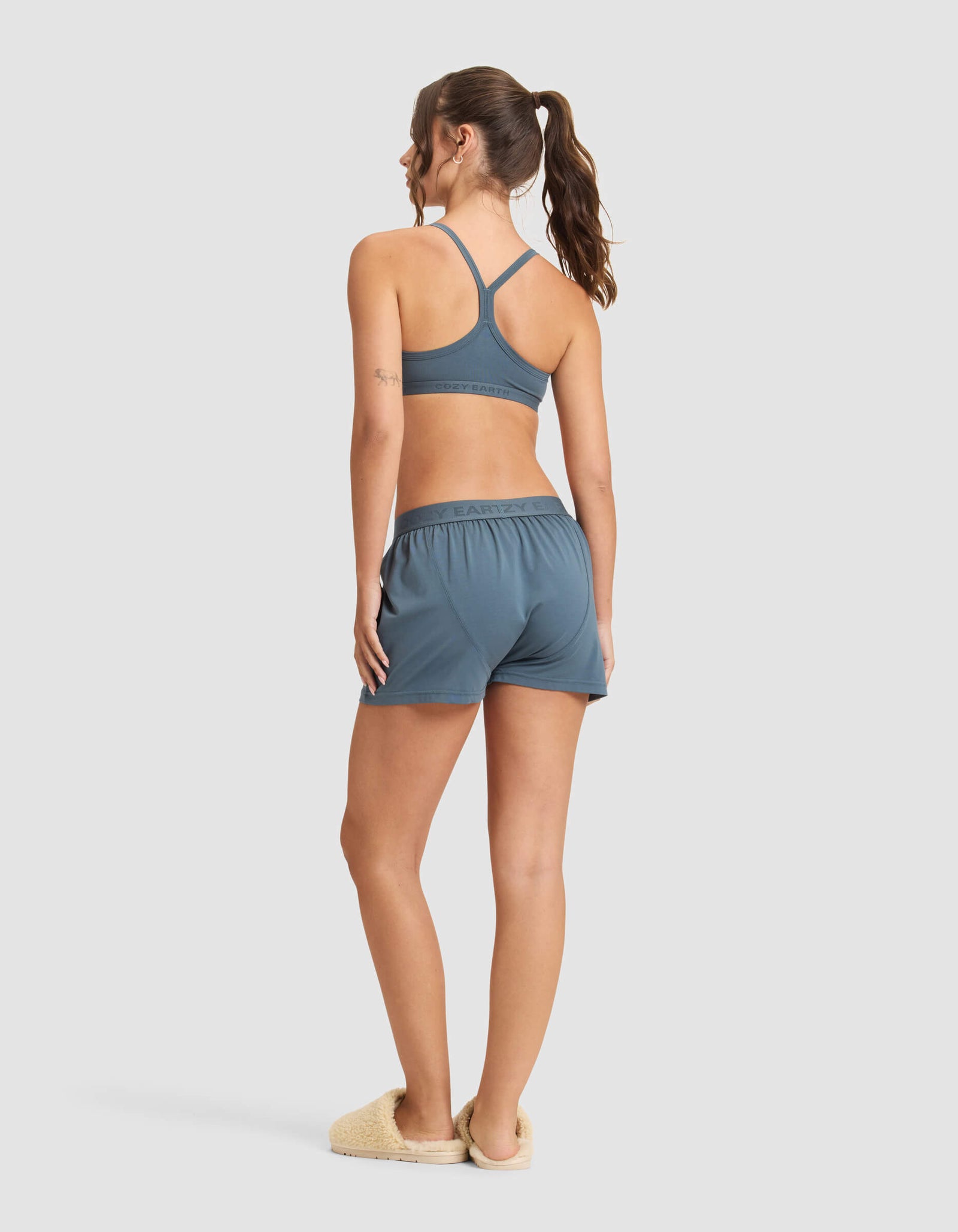 A woman with a ponytail stands facing away, wearing the Cozy Earth Women's First Layer Pajama Soft Bra & Boxer Short Set in blue, paired with beige slippers against a plain light background.