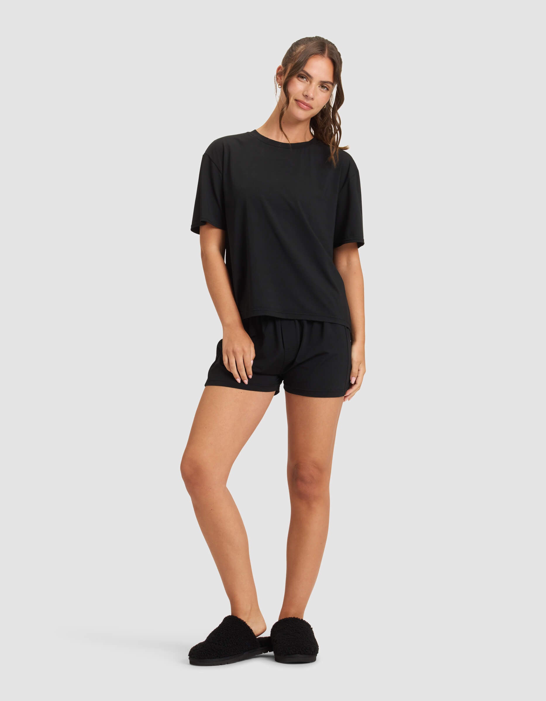 A woman in Cozy Earth's Women's First Layer Pajama Short Sleeve Tee & Boxer Short Set stands against a plain background, smiling softly with her wavy hair pulled back, one hand resting on her thigh.|Color:Jet Black