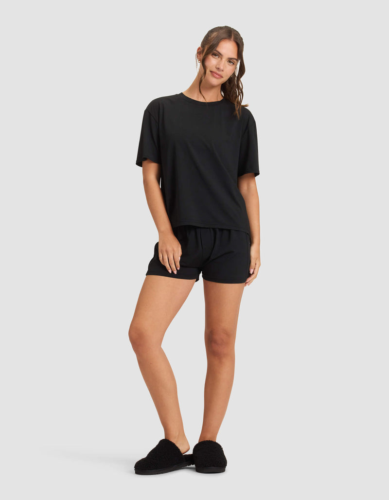 A woman in Cozy Earth's Women's First Layer Pajama Short Sleeve Tee & Boxer Short Set stands against a plain background, smiling softly with her wavy hair pulled back, one hand resting on her thigh.