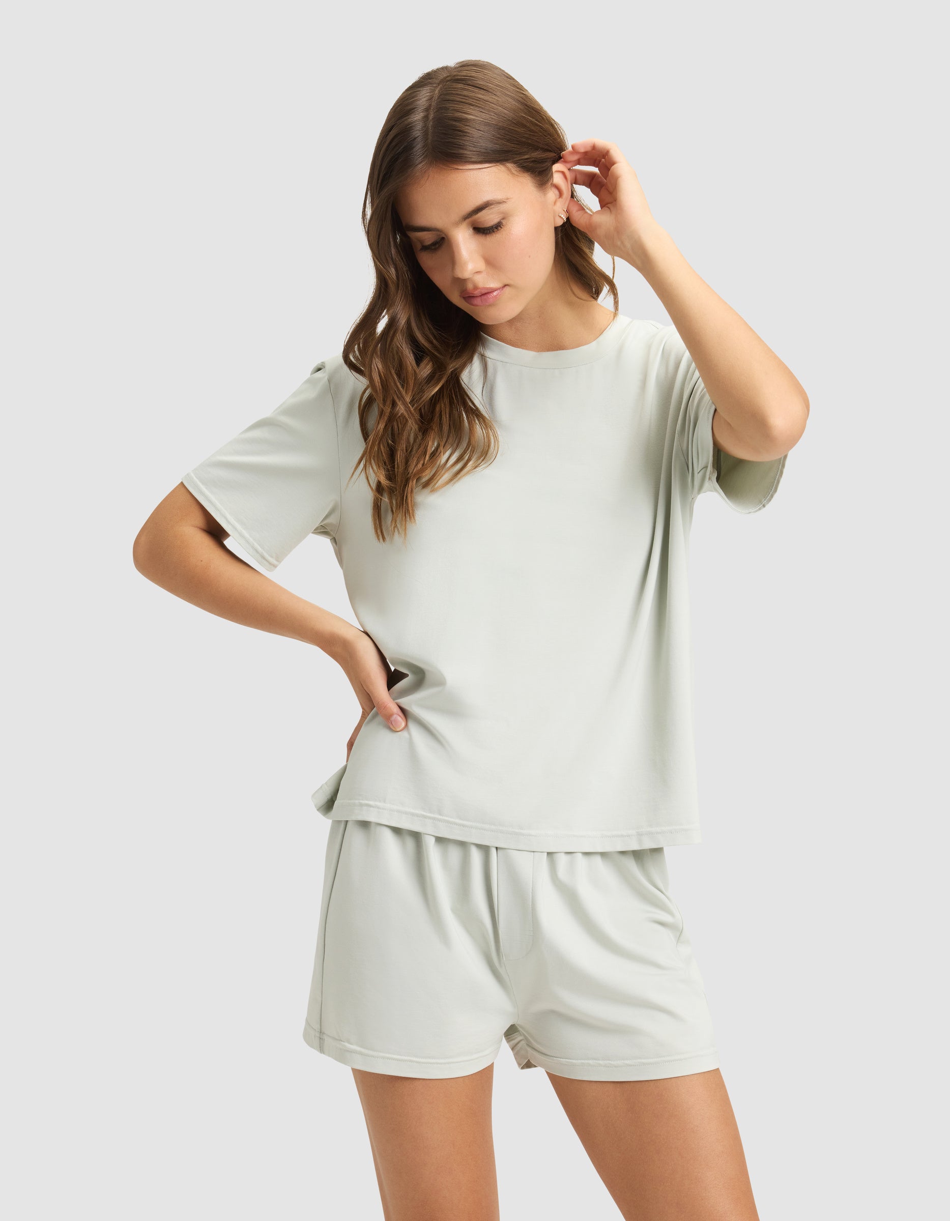 A woman with long brown hair models the Cozy Earth Women's First Layer Pajama Short Sleeve Tee & Boxer Short Set in light gray, standing against a matching background and gently touching her hair while looking down.