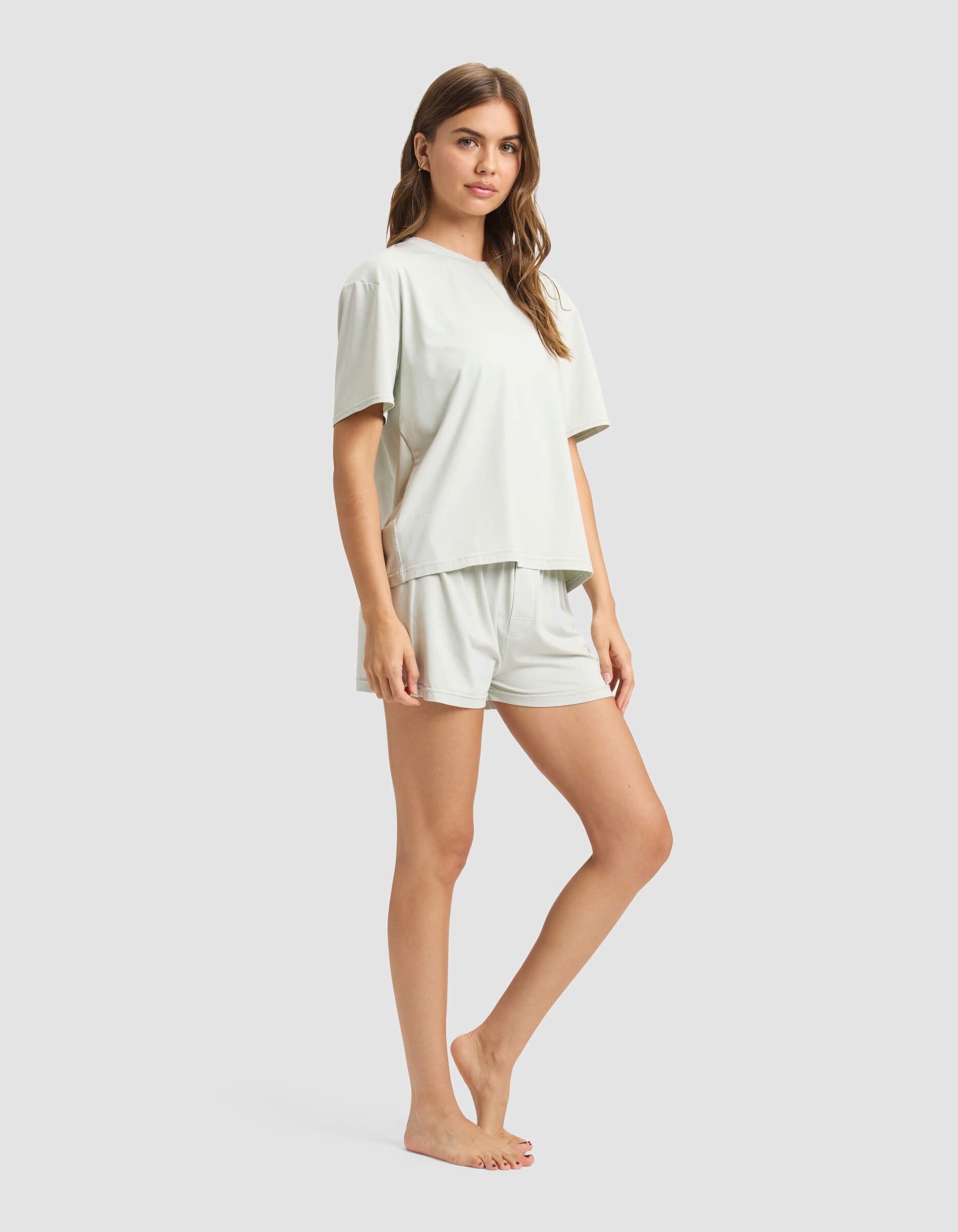 A woman stands barefoot against a plain background, wearing Cozy Earth's Women's First Layer Pajama Short Sleeve Tee & Boxer Short Set in light gray, with her arms relaxed by her sides.