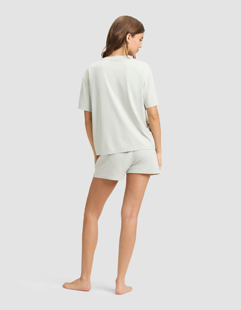 A woman stands barefoot on a light background, facing away, wearing the Cozy Earth Women's First Layer Pajama Short Sleeve Tee & Boxer Short Set in a relaxed fit with her hair down and arms at her sides.