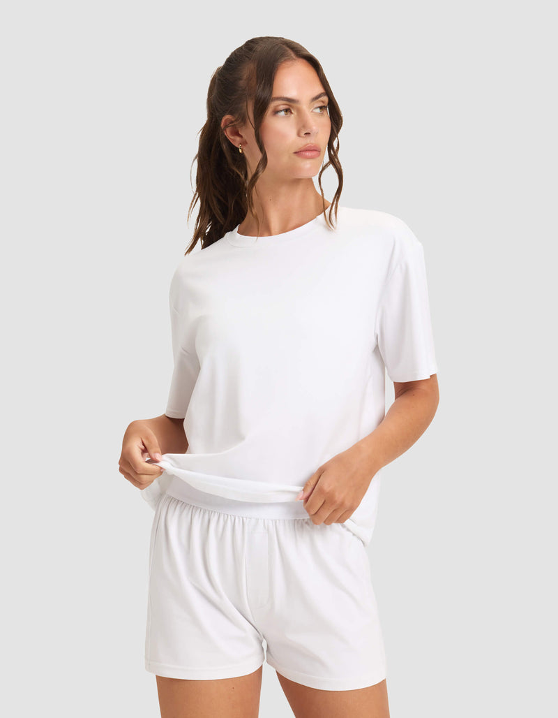 A woman with long brown hair in a ponytail models the Cozy Earth Women's First Layer Pajama Short Sleeve Tee & Boxer Short Set in white, gently adjusting the hem of her shirt against a plain light gray background.