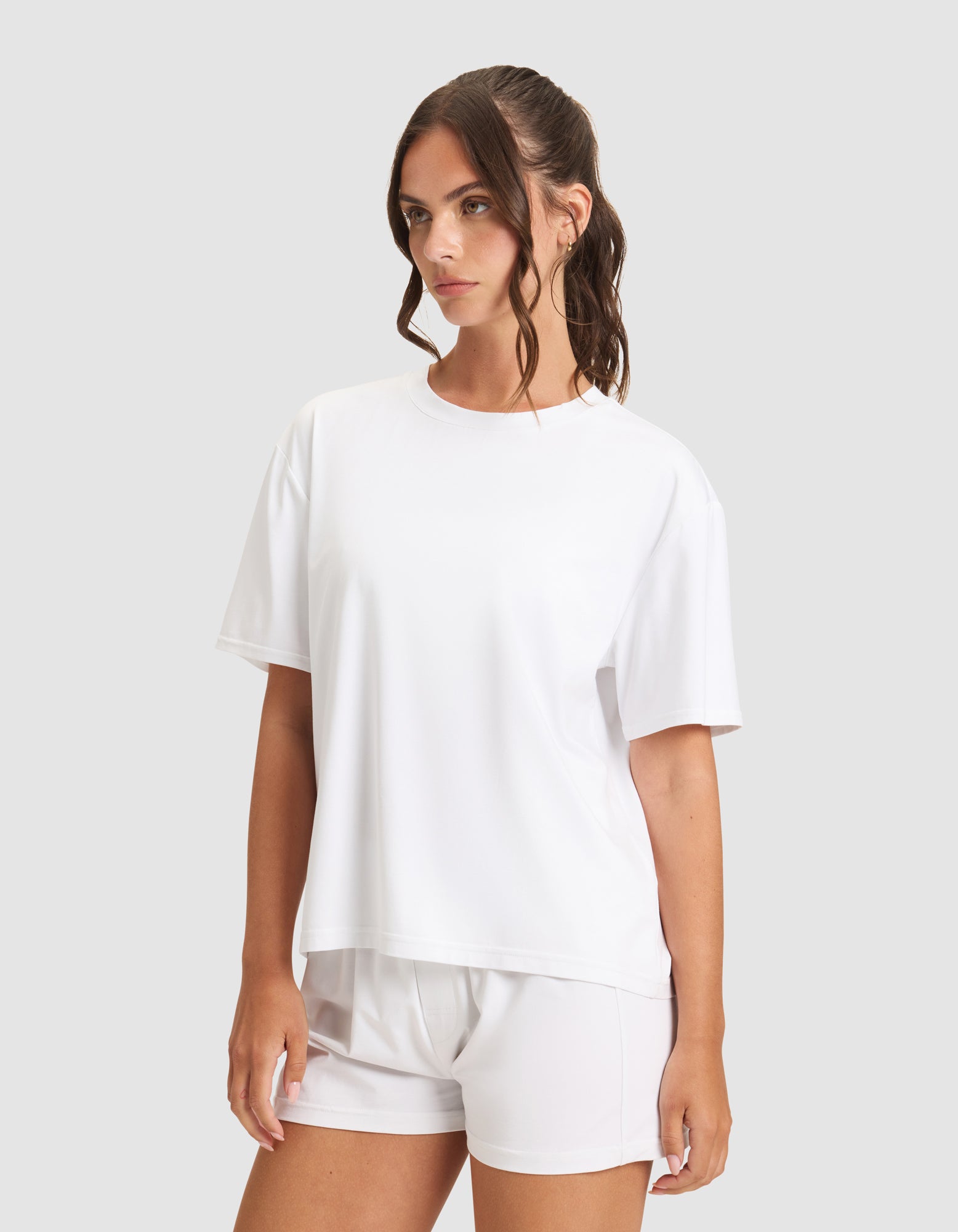 A woman with long brown hair wears the Cozy Earth Women's First Layer Pajama Short Sleeve Tee in white, paired with matching white shorts, standing against a light gray background and looking slightly to the side.