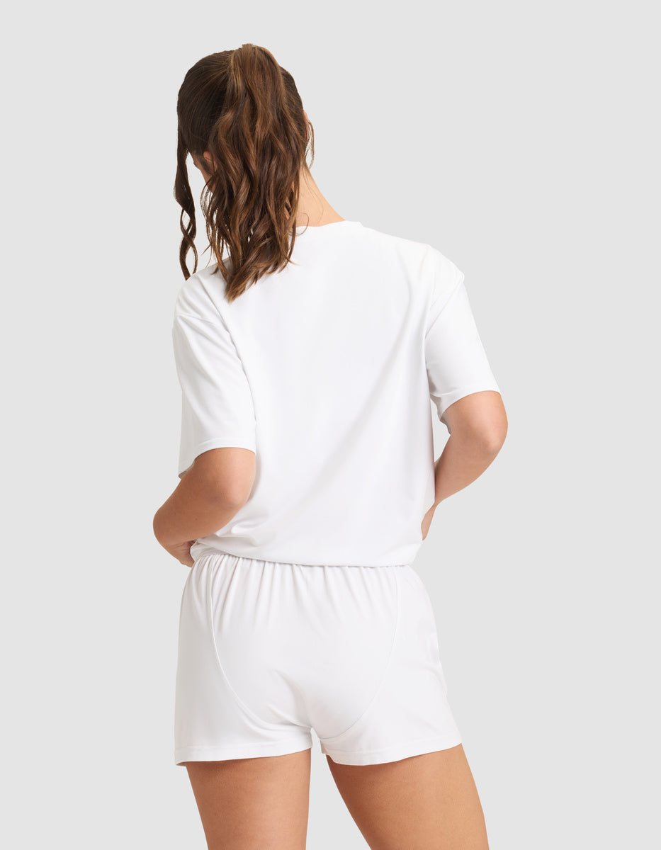 A woman with brown hair in a ponytail faces away, modeling the Cozy Earth Women's First Layer Pajama Short Sleeve Tee & Boxer Short Set in white against a plain light gray background.