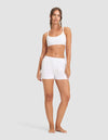 A woman stands barefoot against a plain light gray background, wearing the Cozy Earth Women's First Layer Pajama Soft Bra & Boxer Short Set in white. She faces forward with a neutral expression and loose braided hair.|Color:White