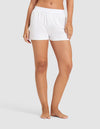 A woman stands barefoot against a plain background, wearing the Cozy Earth Women's First Layer Pajama Boxer Short in white with a matching crop top. Her left hand rests by her side while her right hand gently touches her thigh.|Color:White