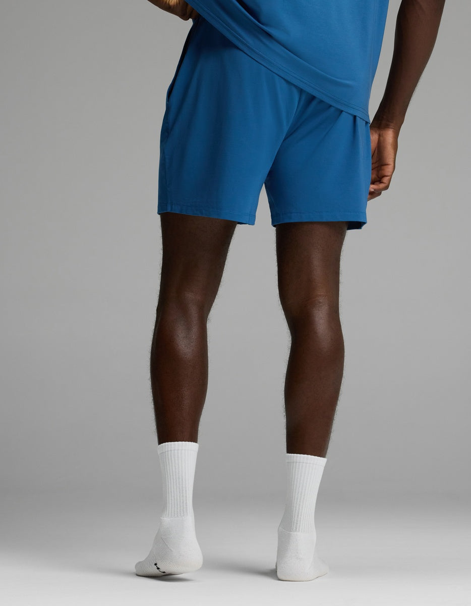 A person stands with their back to the camera, wearing Cozy Earth Men’s First Layer Pajama Shorts in blue, paired with a matching blue shirt and white socks, slightly lifting their shirt. The background is plain and light gray.