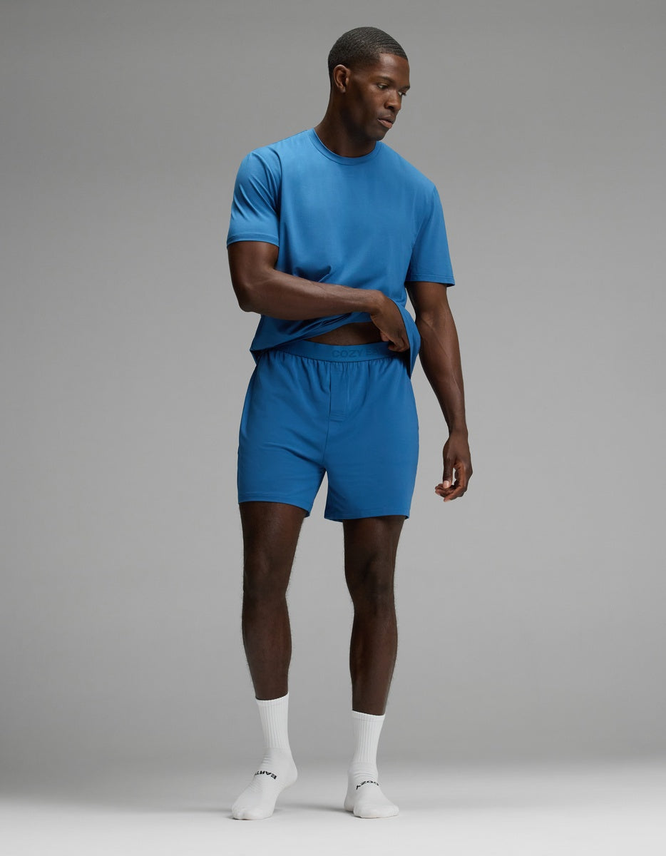 A man stands in a studio wearing Cozy Earth’s Men’s First Layer Pajama Short in blue, paired with a matching t-shirt and white socks. He looks down and points with one hand while his other hand rests on his waist, set against a plain gray background.