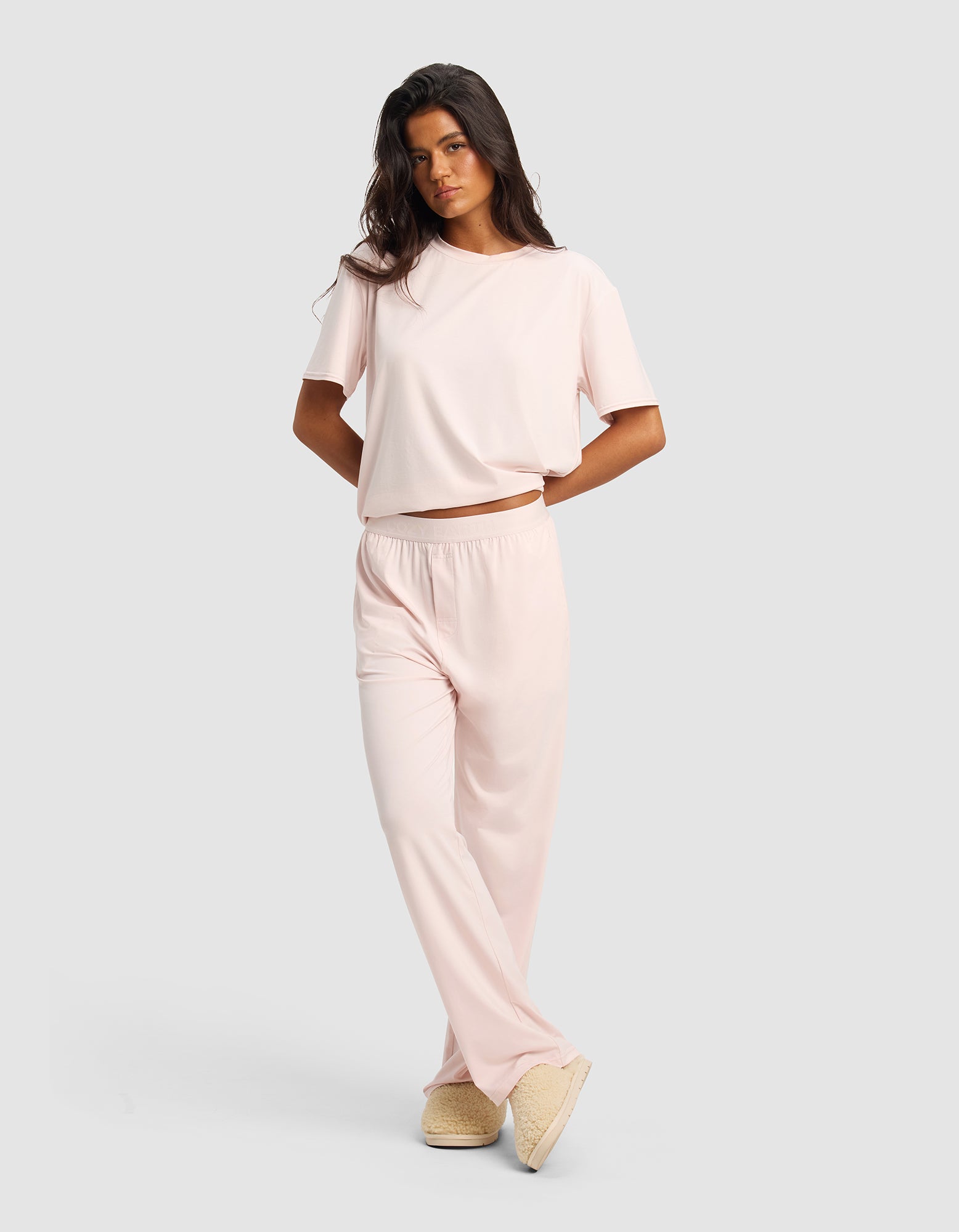 A woman poses against a light gray background wearing the Cozy Earth Women's First Layer Pajama Short Sleeve Tee & Pant Set in light pink with beige slippers, her arms relaxed and legs crossed.|Color:Ballet
