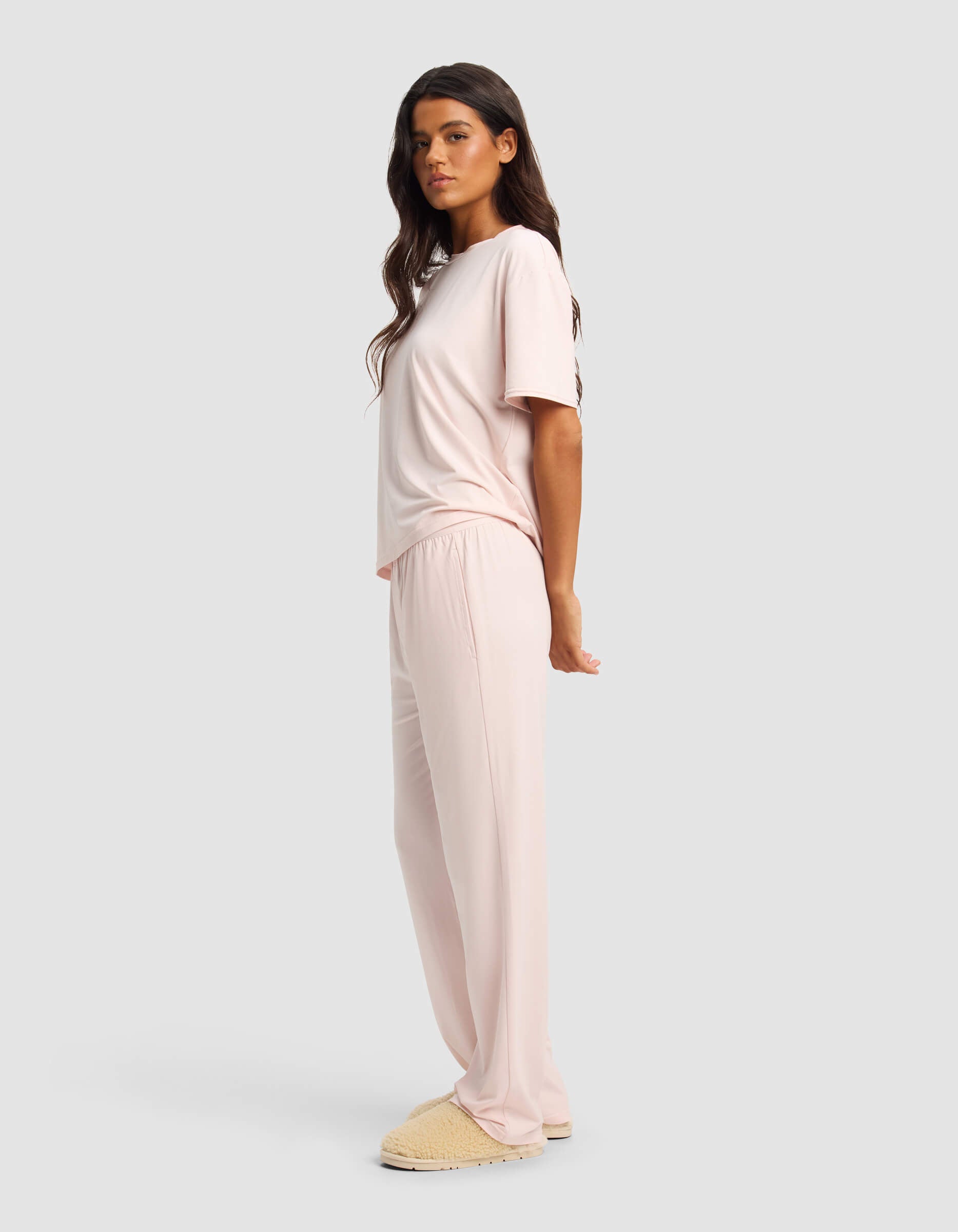 A woman stands sideways in Cozy Earth's Women's First Layer Pajama Short Sleeve Tee & Pant Set in light pink with beige slippers, set against a plain light gray background.|Color:Ballet