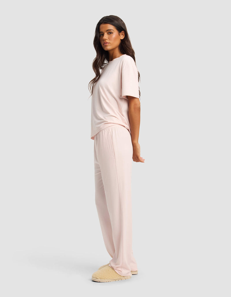 A woman stands sideways in Cozy Earth's Women's First Layer Pajama Short Sleeve Tee & Pant Set in light pink with beige slippers, set against a plain light gray background.