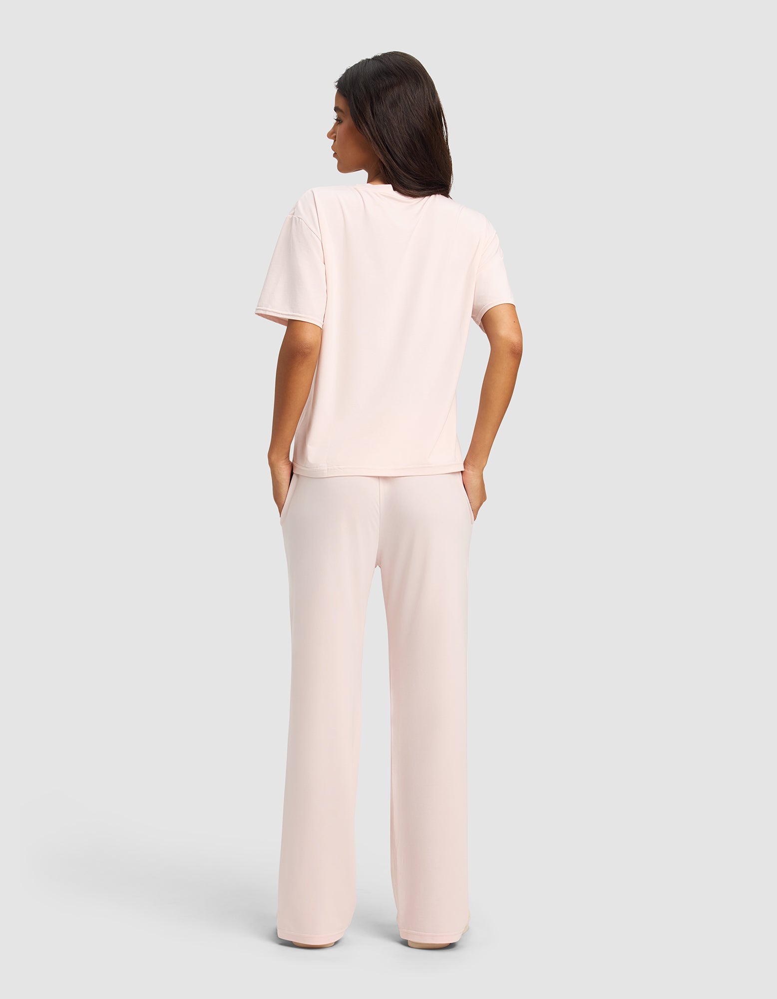 A woman, seen from behind, wears the Cozy Earth Women's First Layer Pajama Short Sleeve Tee & Pant Set in light pink, with hands in her pockets against a plain light gray background.|Color:Ballet