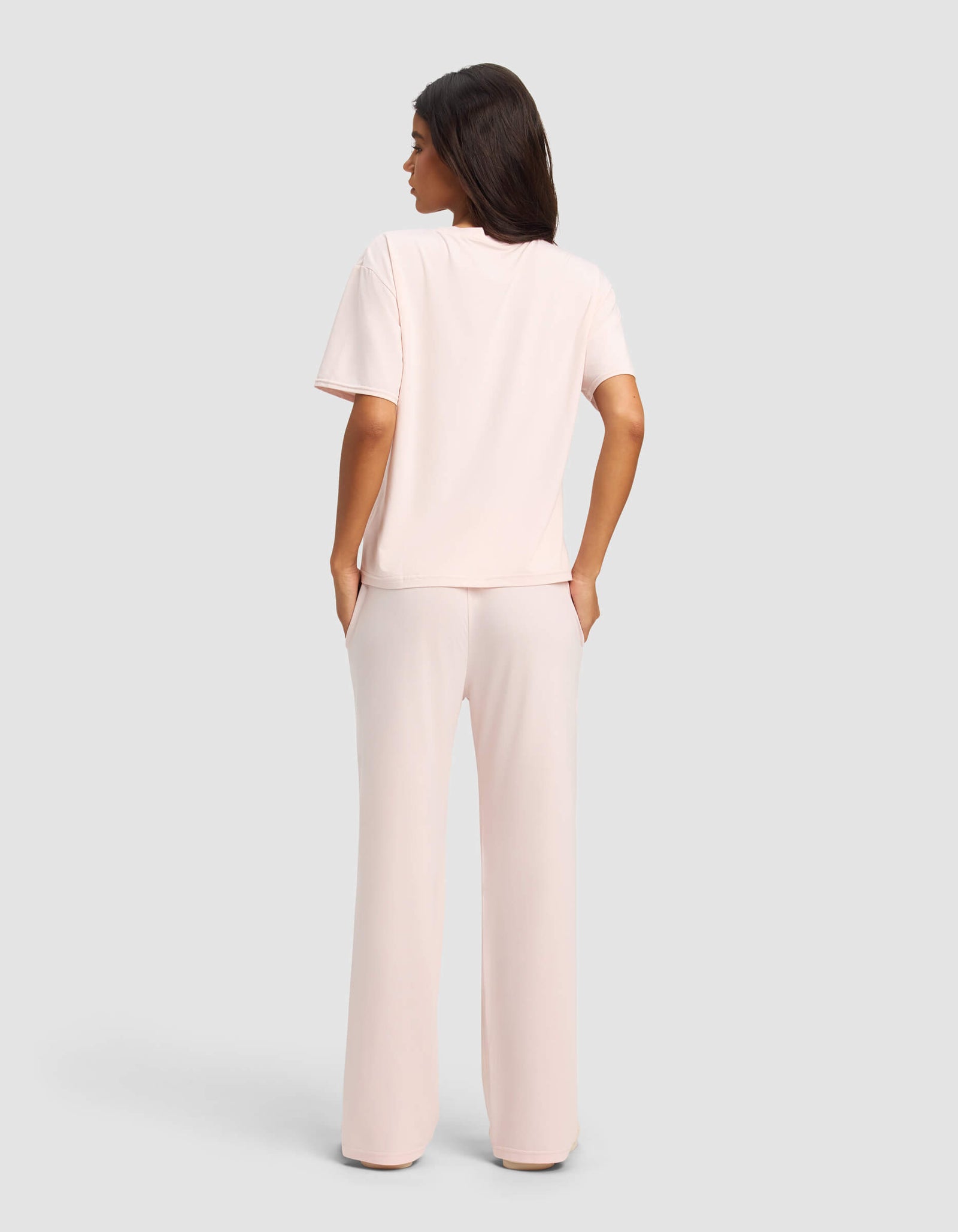 A woman with straight dark hair stands facing away, wearing the Cozy Earth Women's First Layer Pajama Short Sleeve Tee & Pant Set in light pink, with her hands in her pockets, against a plain light gray background.