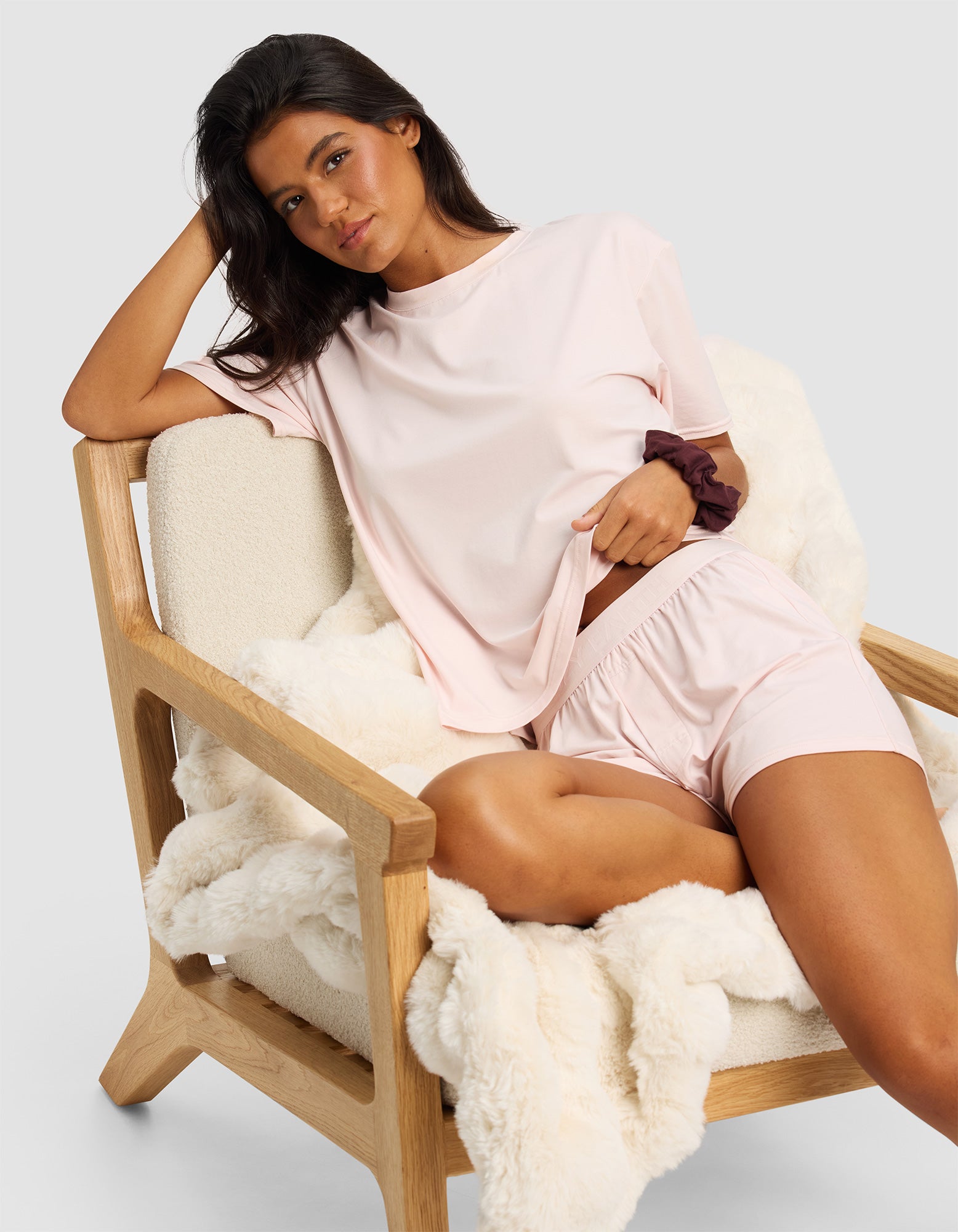 A woman relaxes on a light wooden chair draped with a white blanket, wearing Cozy Earth's Women's First Layer Pajama Short Sleeve Tee & Boxer Short Set, showcasing comfort and style in pale pink.