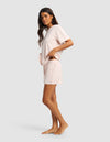 A woman with long dark hair wears the Cozy Earth Women's First Layer Pajama Short Sleeve Tee & Boxer Short Set in light pink, standing barefoot against a plain light gray background and looking slightly toward the camera.|Color:Ballet