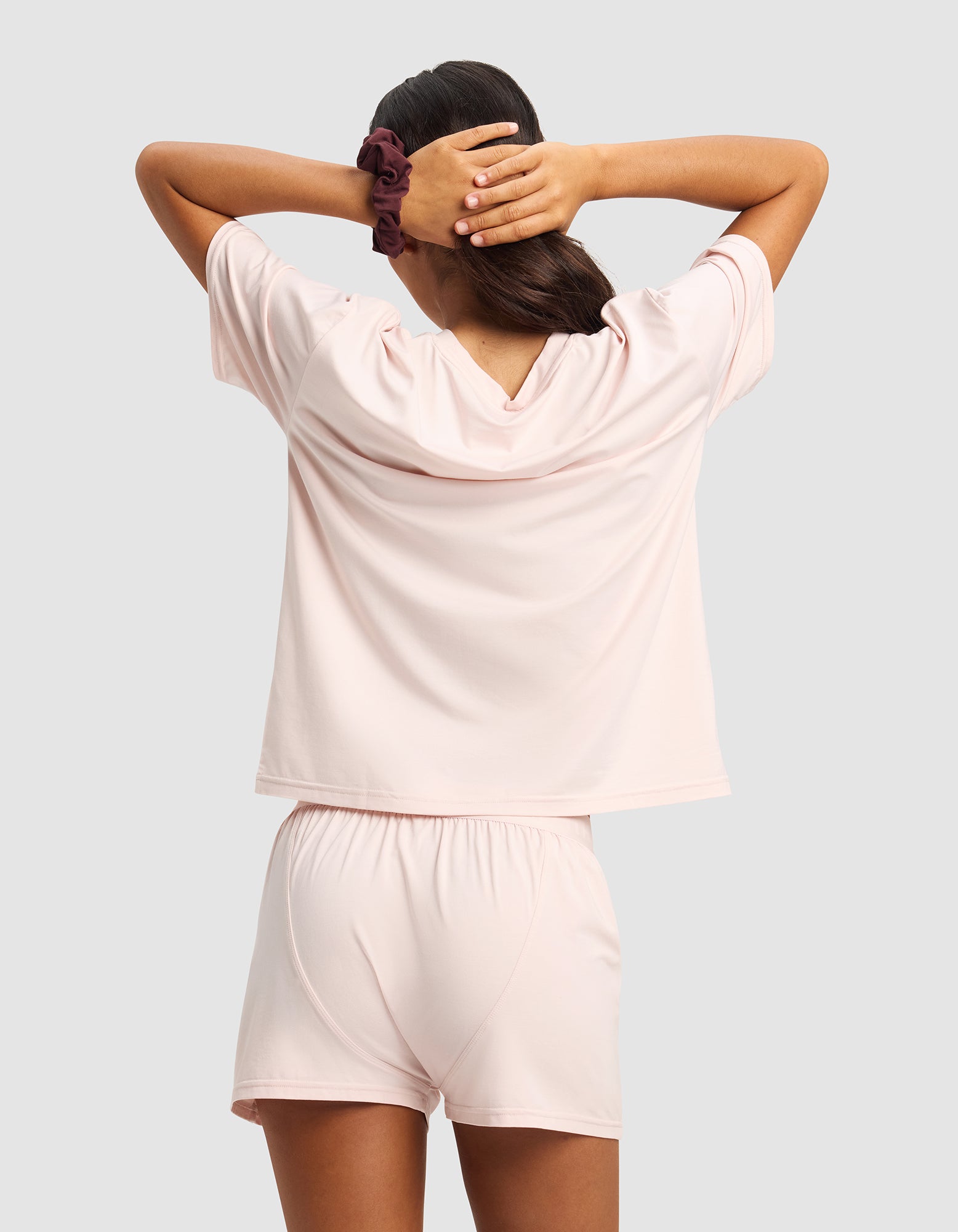 A woman with long dark hair wears the Cozy Earth Women's First Layer Pajama Short Sleeve Tee & Boxer Short Set in light pink, standing with her back to the camera and hands behind her head, a dark scrunchie on one wrist.