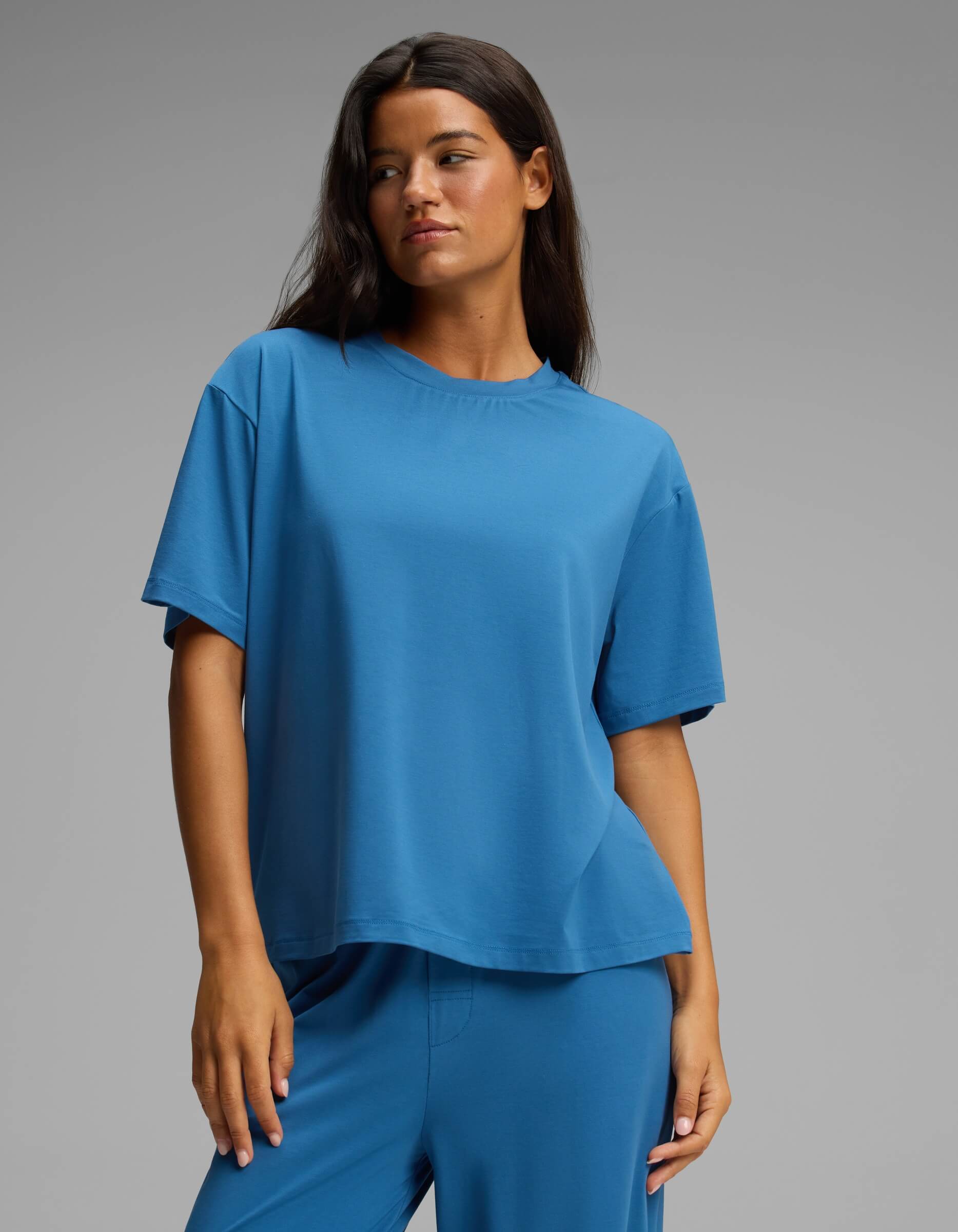 A woman with long dark hair wears the Cozy Earth Women’s First Layer Pajama Short Sleeve Tee in blue, paired with matching blue pants. She stands against a plain gray background, looking slightly to the side with a neutral expression.