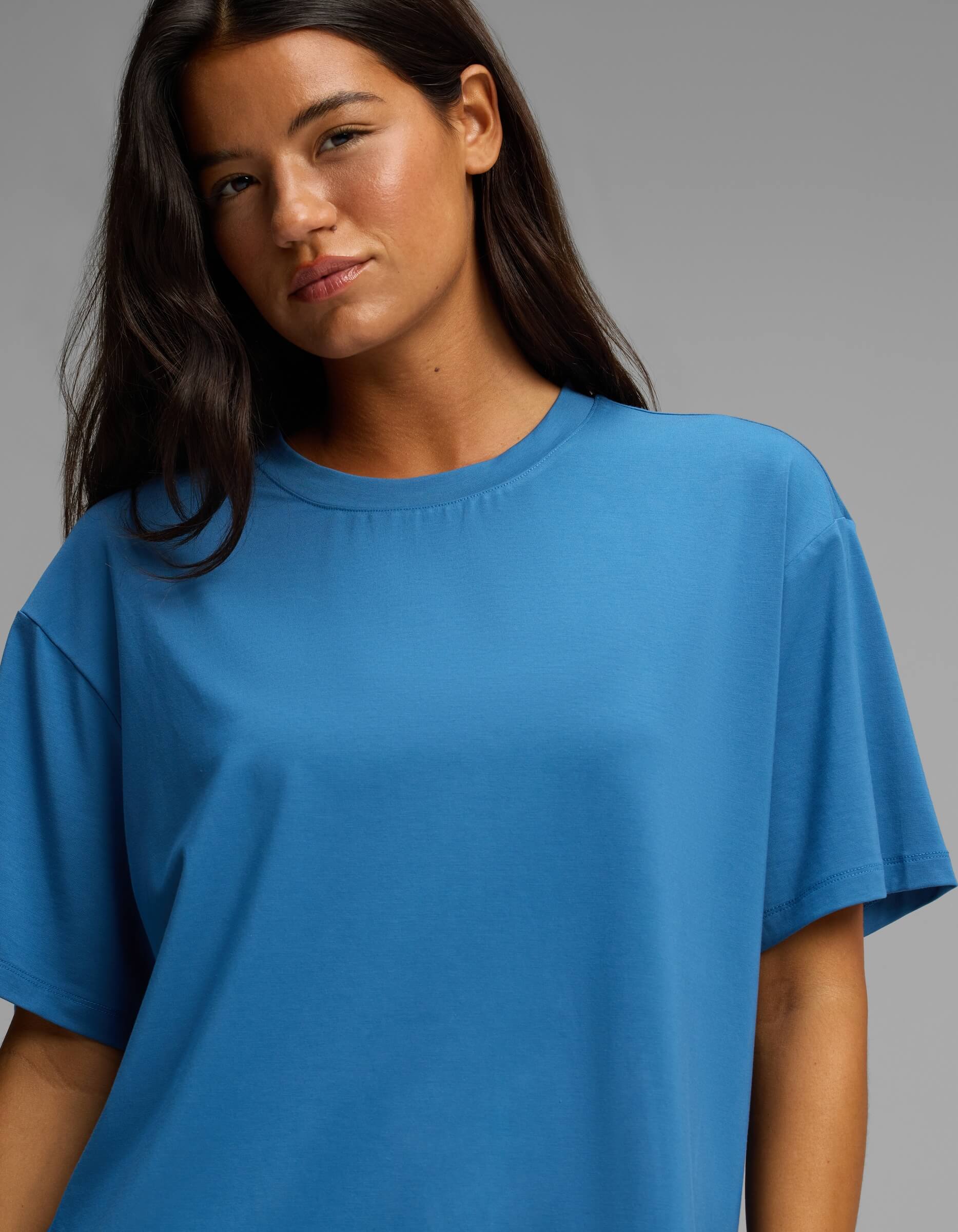 A woman with long dark hair wears the Cozy Earth Women’s First Layer Pajama Short Sleeve Tee in blue, standing against a plain gray background and looking slightly towards the camera with a neutral expression.
