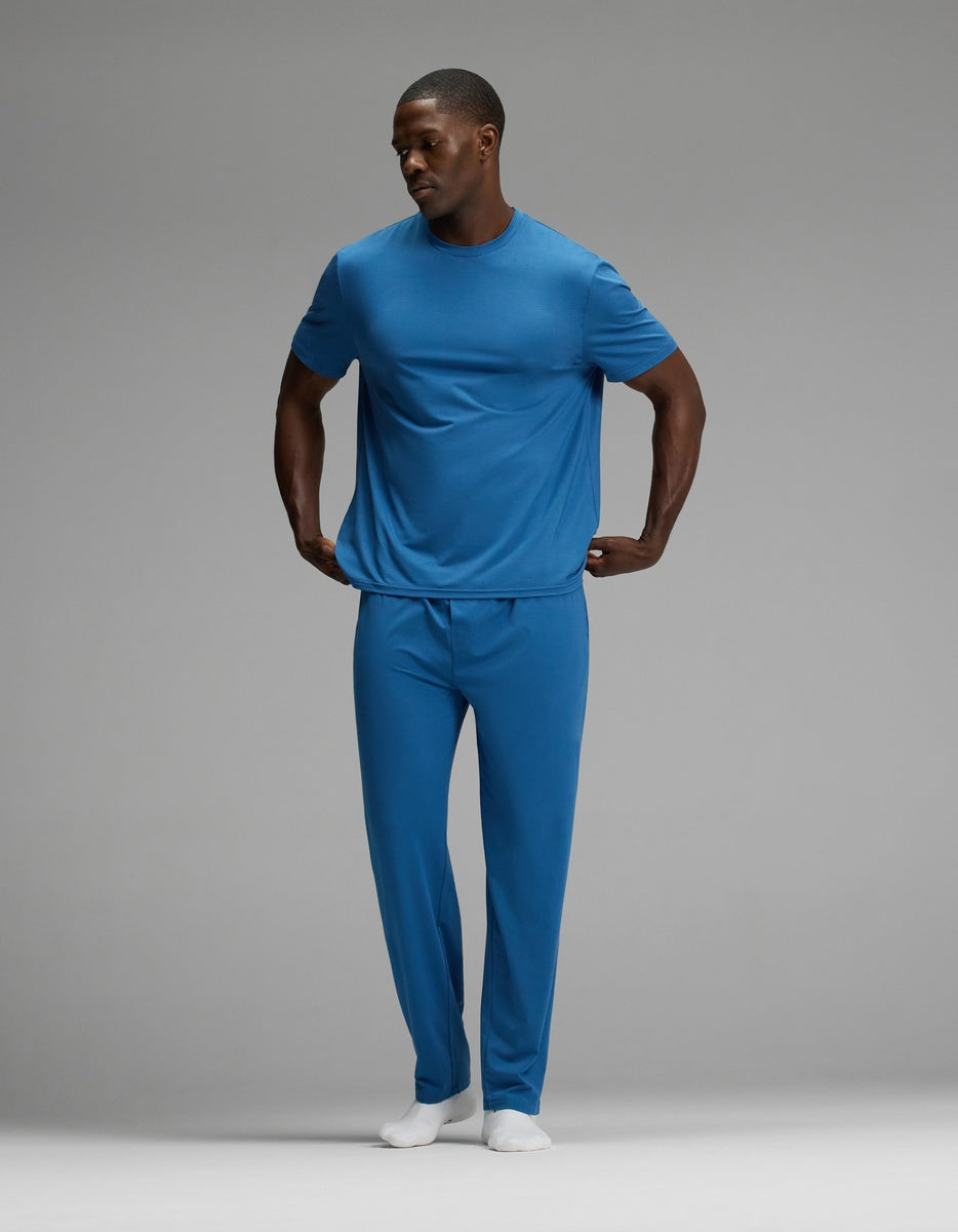 A man stands with his hands on his hips, wearing the Cozy Earth Men’s First Layer Pajama Short Sleeve Tee and matching pants, along with white socks, against a plain gray background.