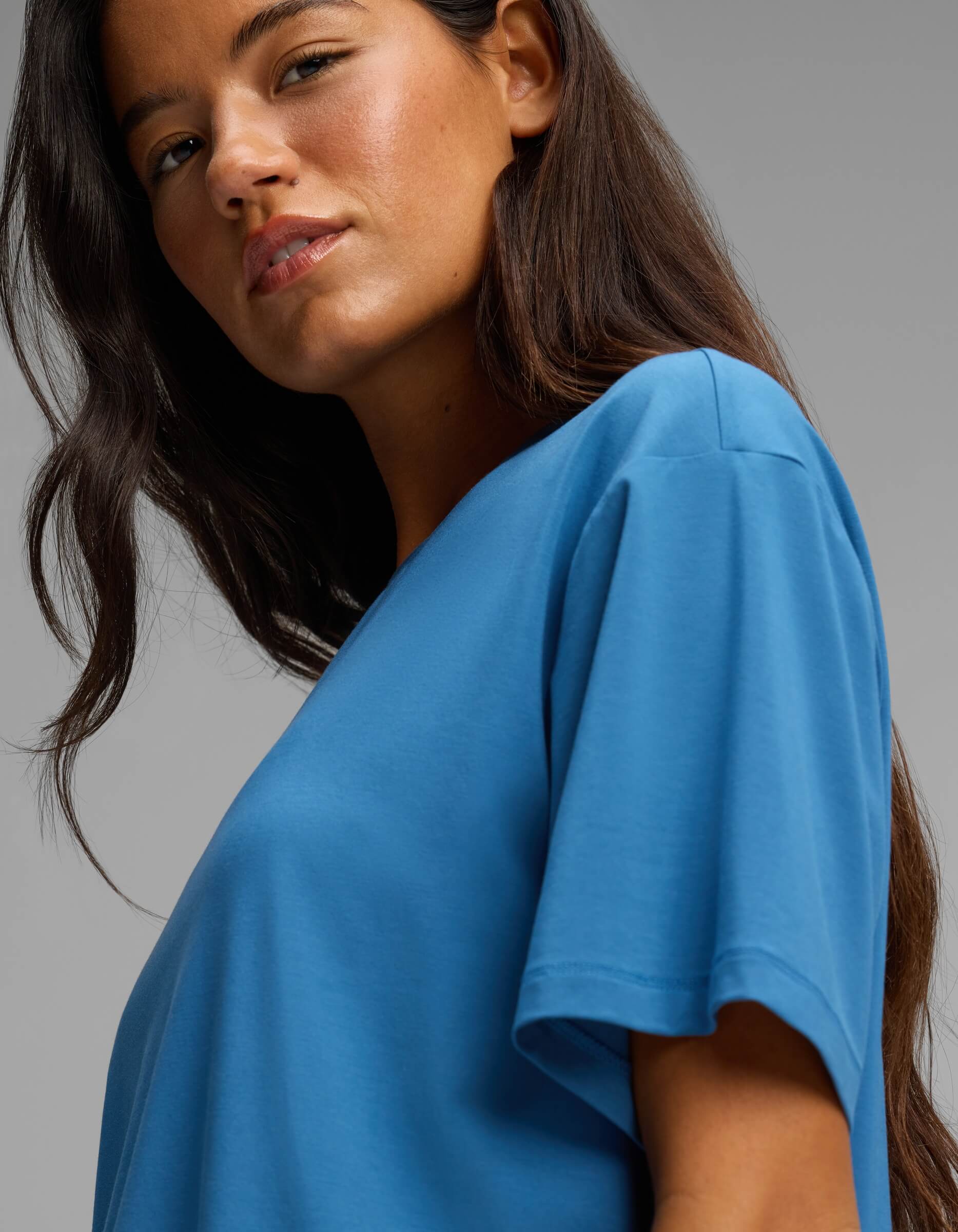 A woman with long brown hair, wearing the Cozy Earth Women’s First Layer Pajama Short Sleeve Tee in blue, looks slightly over her shoulder against a neutral gray background.