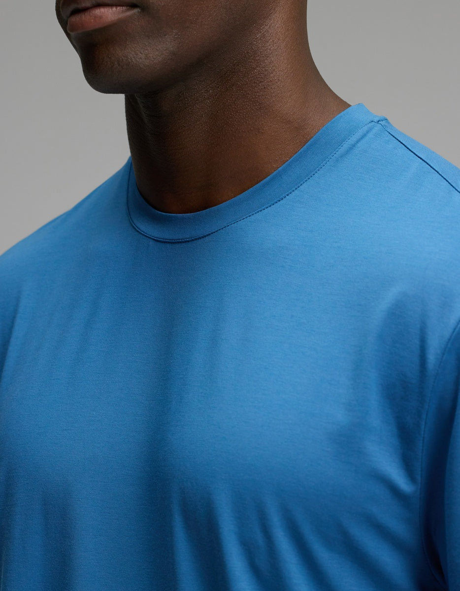 Close-up of a person’s upper torso and neck wearing the Cozy Earth Men’s First Layer Pajama Short Sleeve Tee in a plain blue color against a neutral background. The person’s head is partially visible, with facial features not fully shown.