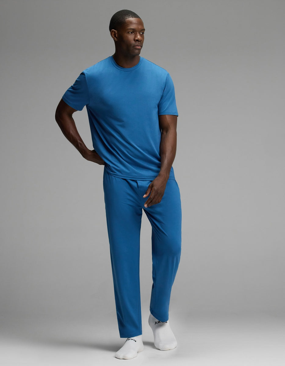 A man stands against a plain gray background wearing the Cozy Earth Men’s First Layer Pajama Short Sleeve Tee in blue, paired with matching blue pants and white socks. He has one hand on his hip and the other resting by his thigh.