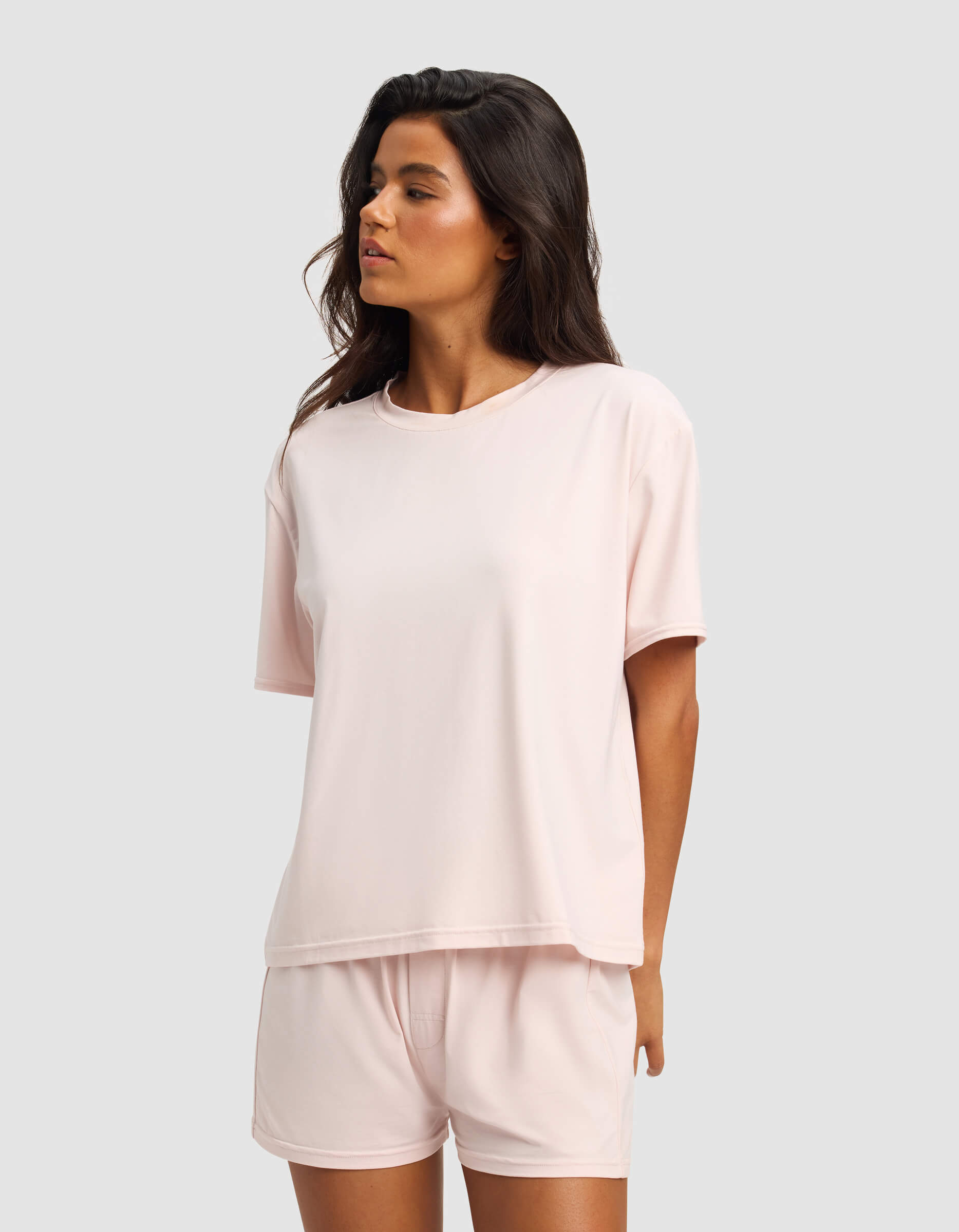 A woman with long dark hair wears the Cozy Earth Women's First Layer Pajama Short Sleeve Tee and matching shorts, standing against a plain light gray background and looking to the side.|Color:Ballet