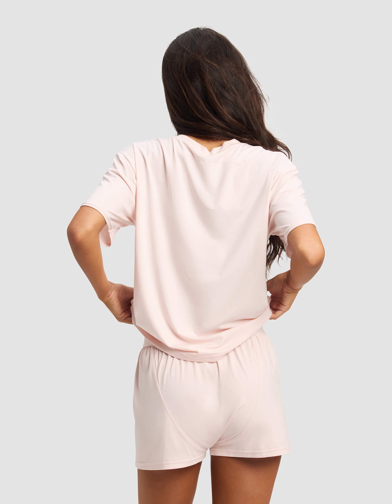 A woman with long brown hair faces away from the camera, wearing Cozy Earth’s Women's First Layer Pajama Short Sleeve Tee and matching shorts in pale pink. She stands with hands on hips against a plain light gray background.