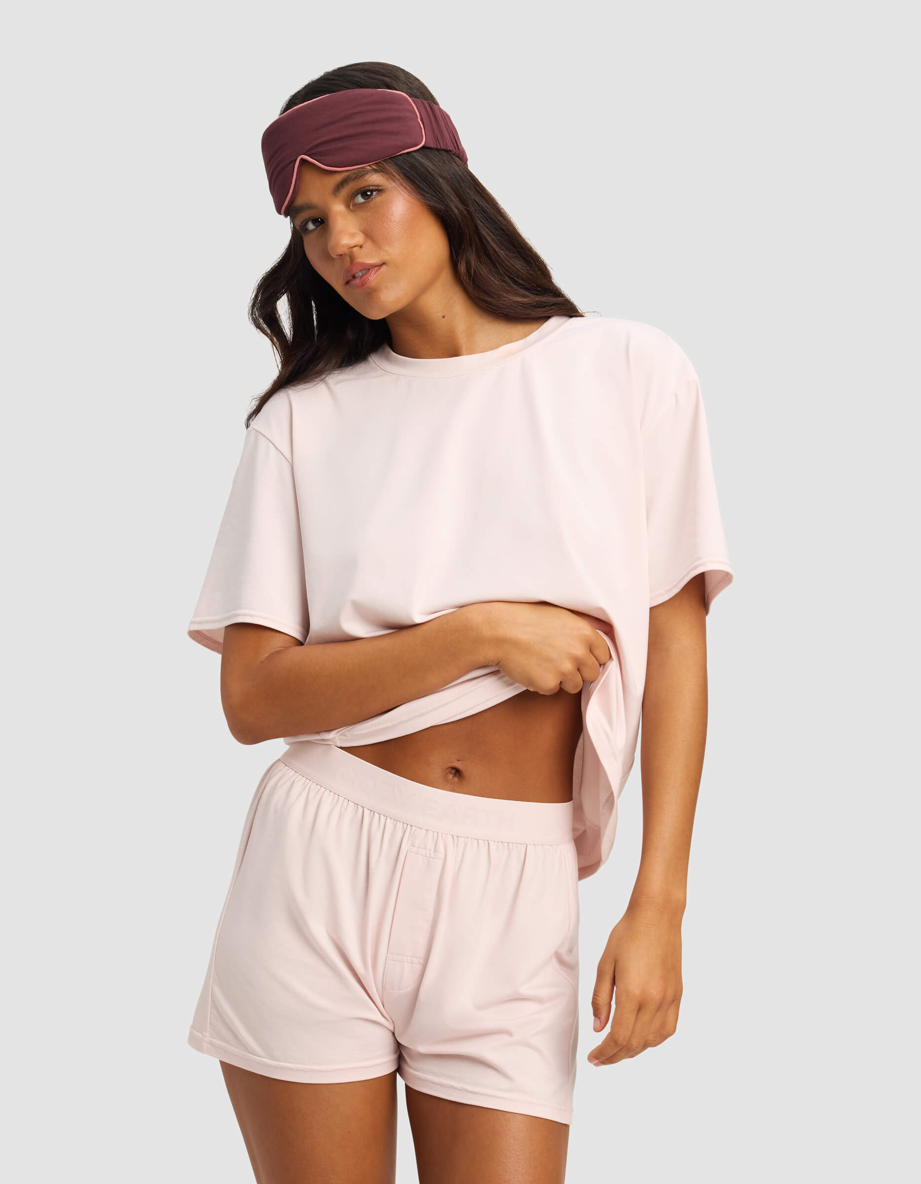 A woman in Cozy Earth's Women's First Layer Pajama Short Sleeve Tee poses against a light gray background, lifting her loose pink top to show matching shorts and wearing a dark pink sleep mask.|Color:Ballet