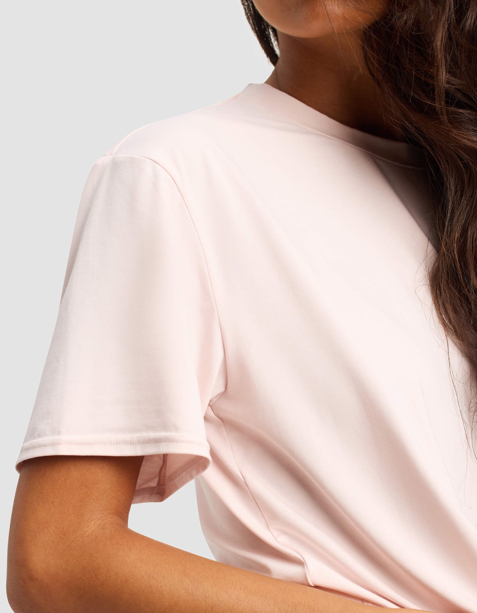 A close-up of a woman wearing the Cozy Earth Women's First Layer Pajama Short Sleeve Tee in light pink, with wavy brown hair over her shoulder, set against a plain light grey background.