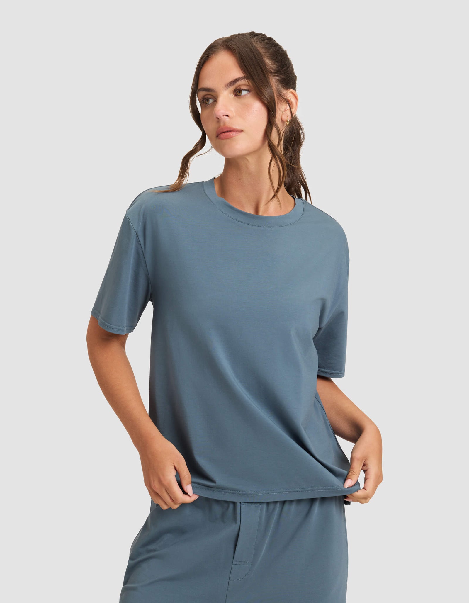 A woman wearing Cozy Earth's Women's First Layer Pajama Short Sleeve Tee and matching pants stands against a plain light gray background, looking to the side.