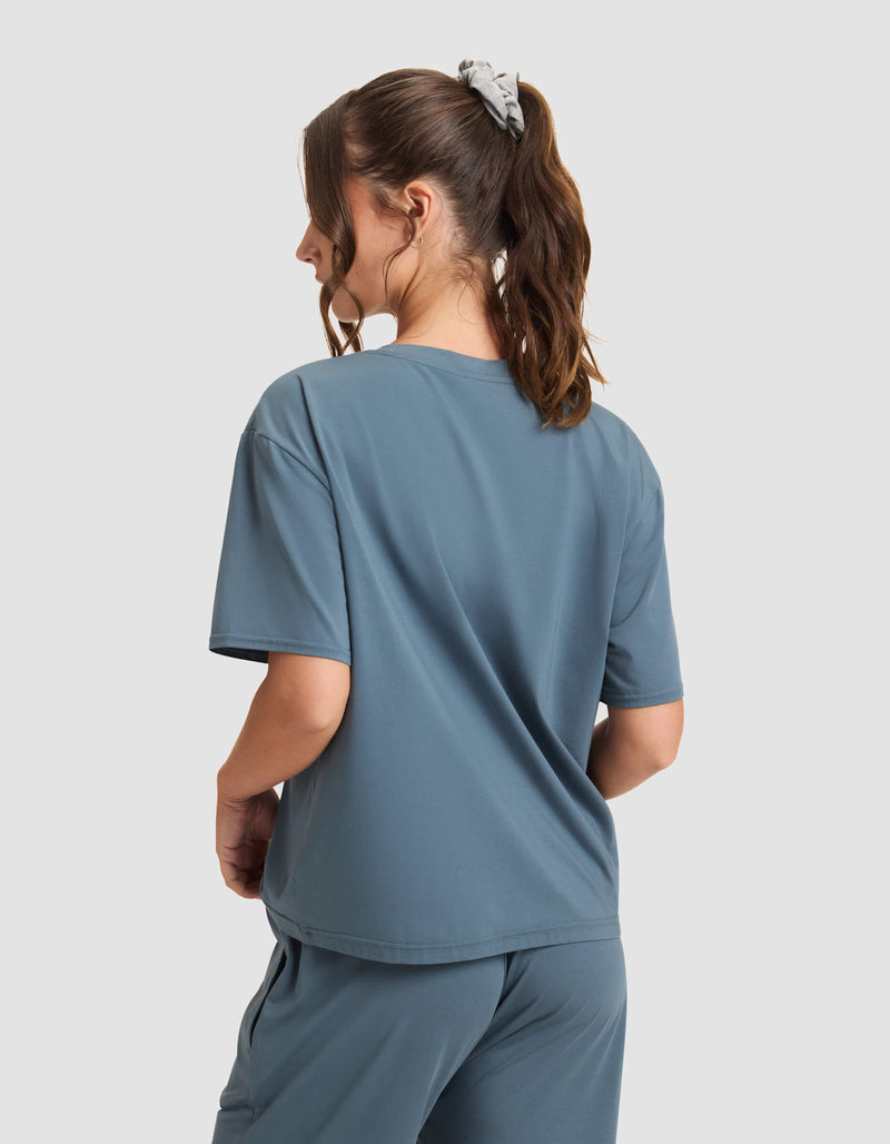A woman with a brown ponytail and gray scrunchie is seen from behind, wearing Cozy Earth’s Women's First Layer Pajama Short Sleeve Tee in blue pants and top, standing against a plain light gray background.