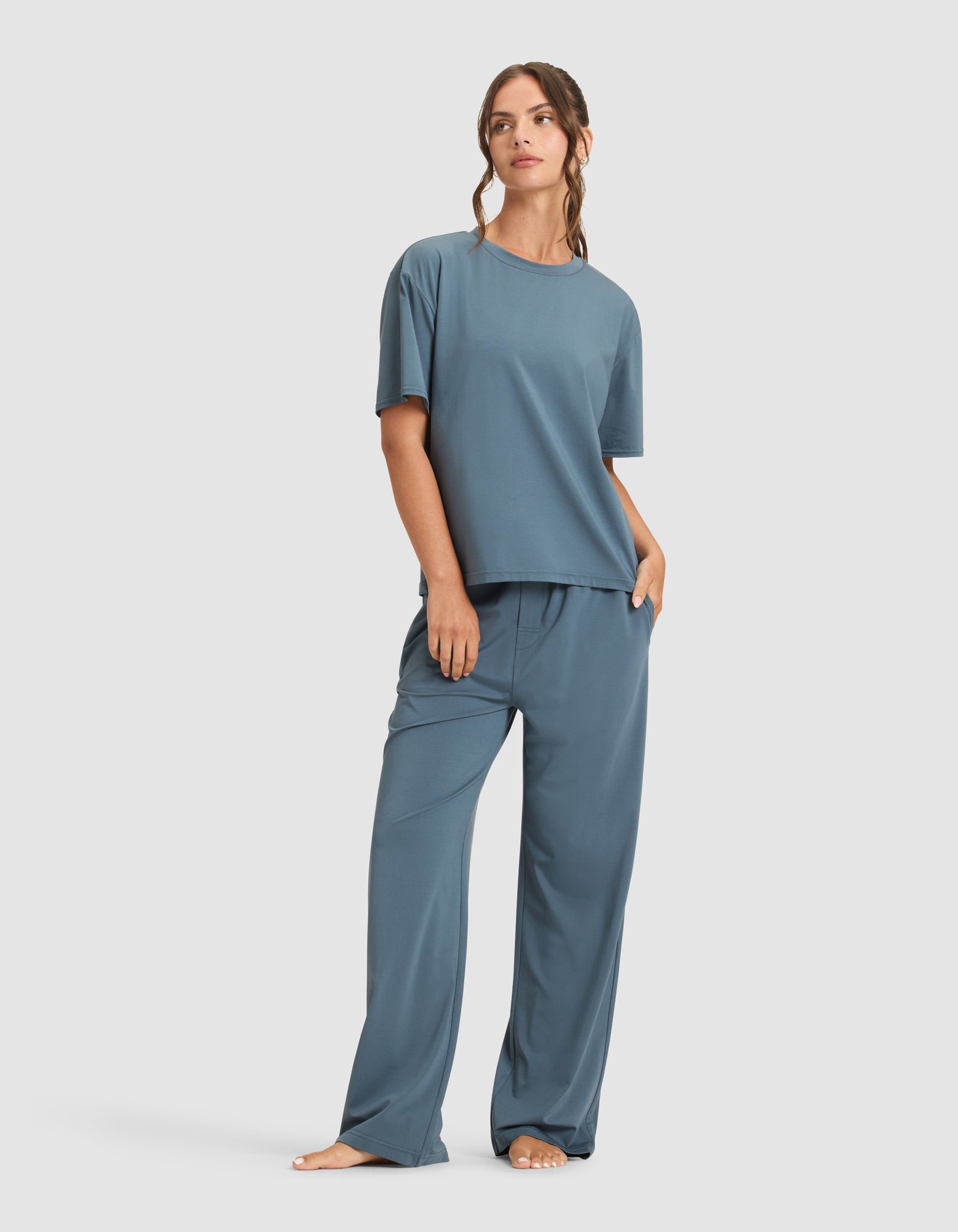 A woman stands barefoot against a plain background, wearing Cozy Earth's Women's First Layer Pajama Short Sleeve Tee and matching wide-leg pants. She looks to the side with one hand in her pocket.
