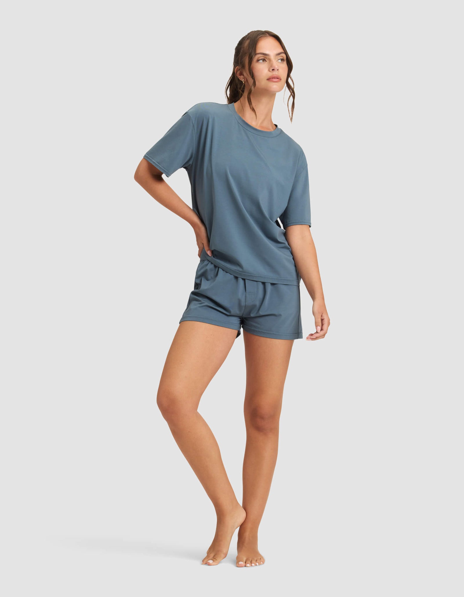 A woman poses barefoot against a plain background, wearing Cozy Earth's Women's First Layer Pajama Short Sleeve Tee in blue with matching shorts, standing casually with one leg slightly bent.