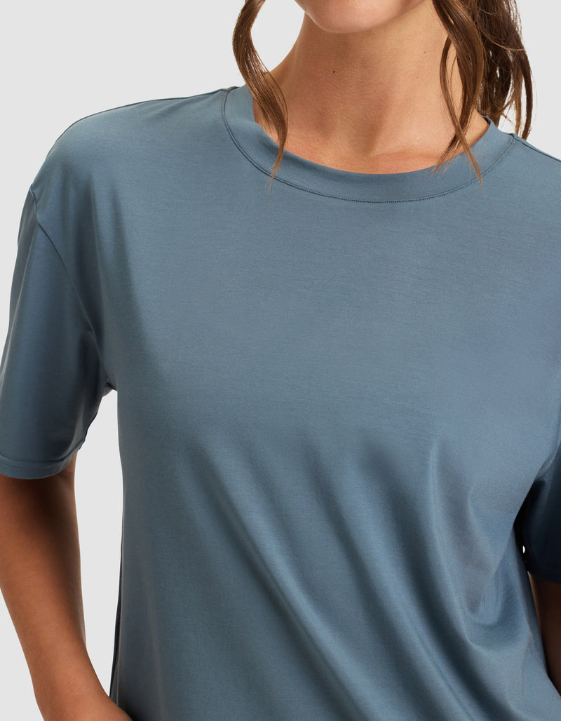 A woman stands with one hand on her hip, wearing the Cozy Earth Women's First Layer Pajama Short Sleeve Tee in light blue. Her head is partially visible, and her brown hair is partially tied back.