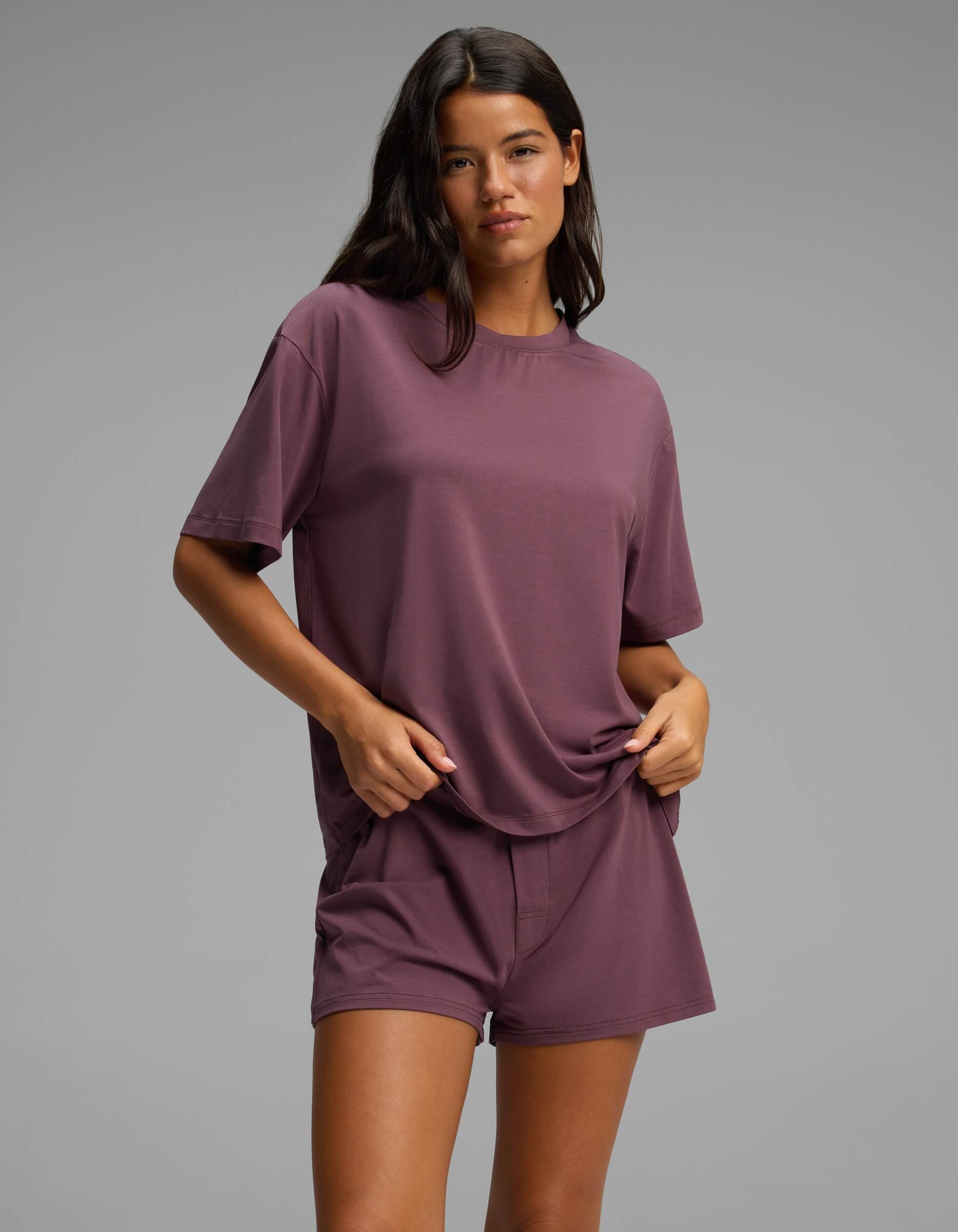 A woman stands against a plain gray background, wearing the Cozy Earth Women’s First Layer Pajama Short Sleeve Tee in mauve with matching shorts, one hand holding her shirt and the other in her pocket.