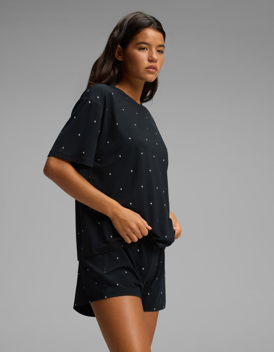 A person stands sideways against a plain gray background, wearing the Cozy Earth Women’s First Layer Pajama Short Sleeve Tee & Boxer Short Set in black with small white dots.