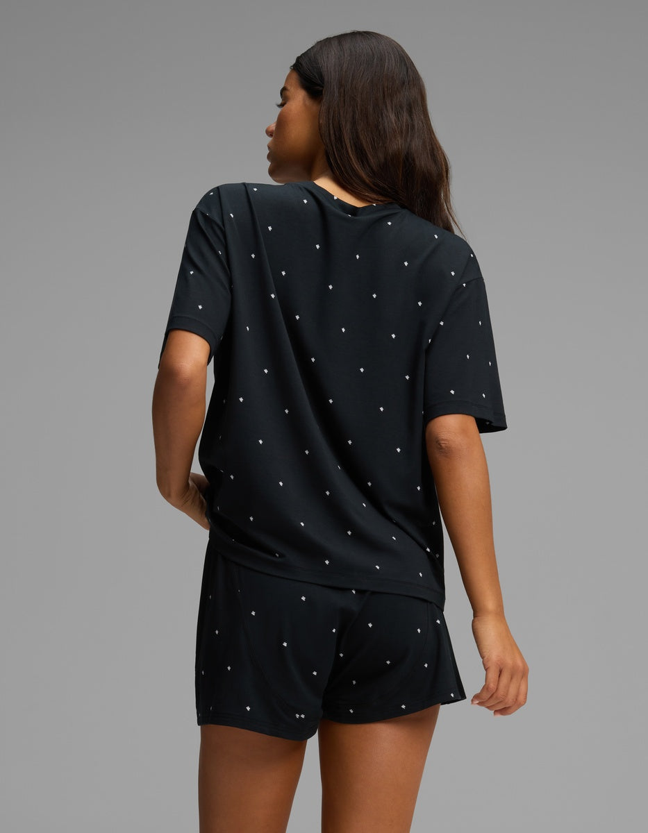 A person stands against a plain background, seen from behind with one arm bent, wearing the Cozy Earth Women’s First Layer Pajama Short Sleeve Tee & Boxer Short Set in black with small white polka dots.