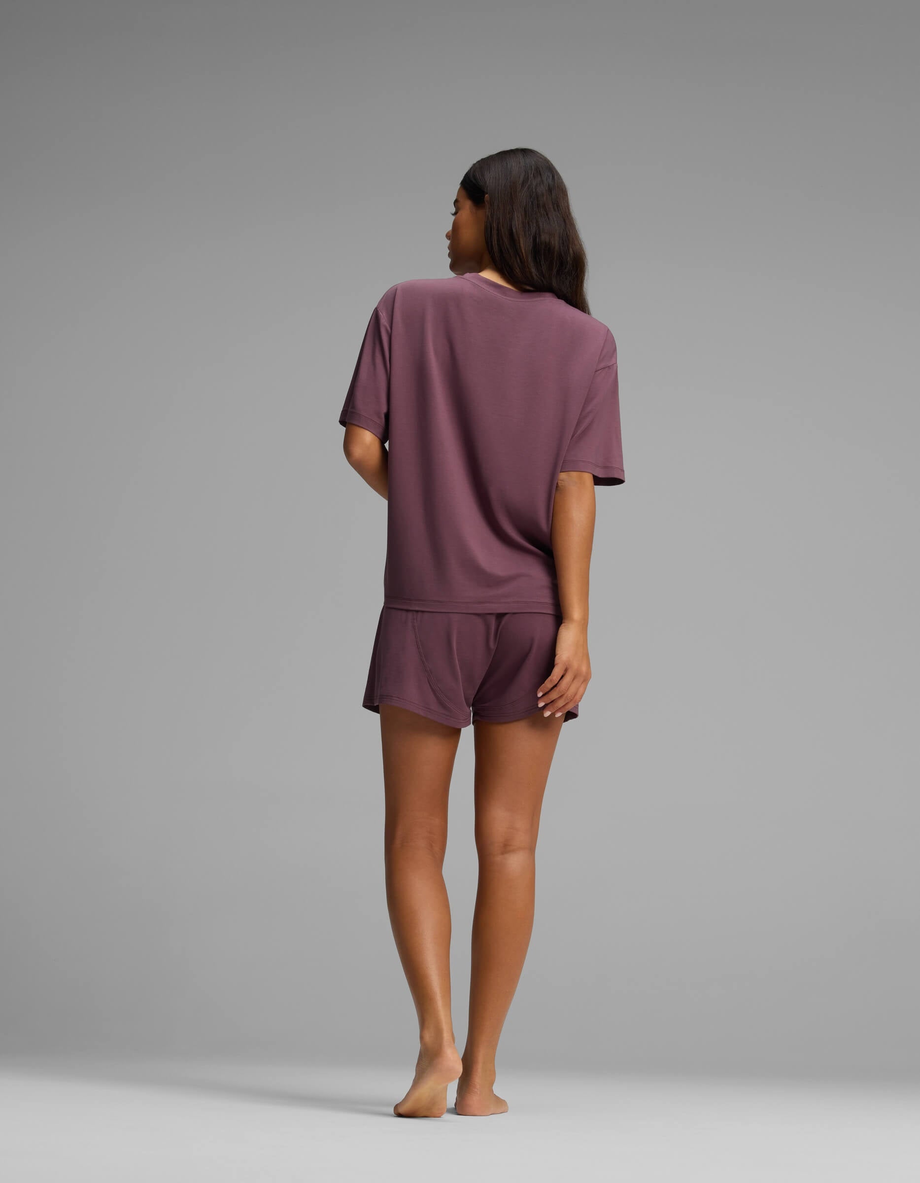A woman stands barefoot, facing away, wearing the Cozy Earth Women’s First Layer Pajama Short Sleeve Tee & Boxer Short Set in mauve against a plain gray background.