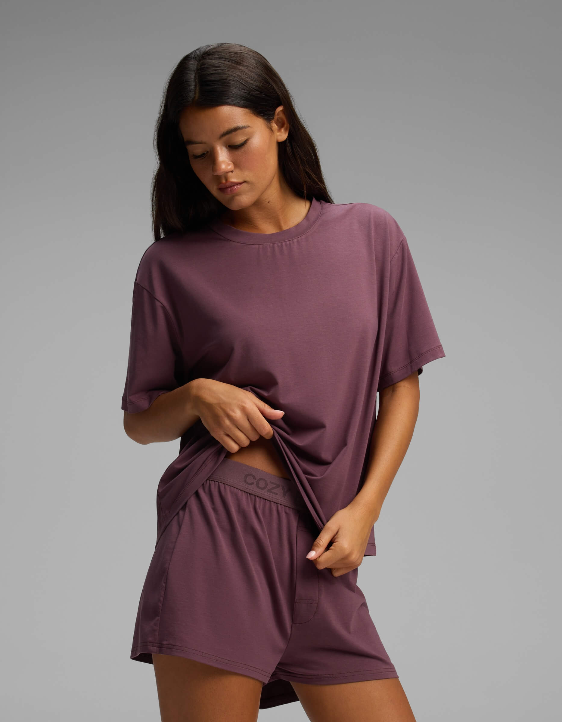 A woman stands against a plain background, slightly lifting the hem of her shirt to reveal the waistband of her shorts, while wearing the Cozy Earth Women’s First Layer Pajama Short Sleeve Tee & Boxer Short Set in a matching mauve color.|Color:Fig