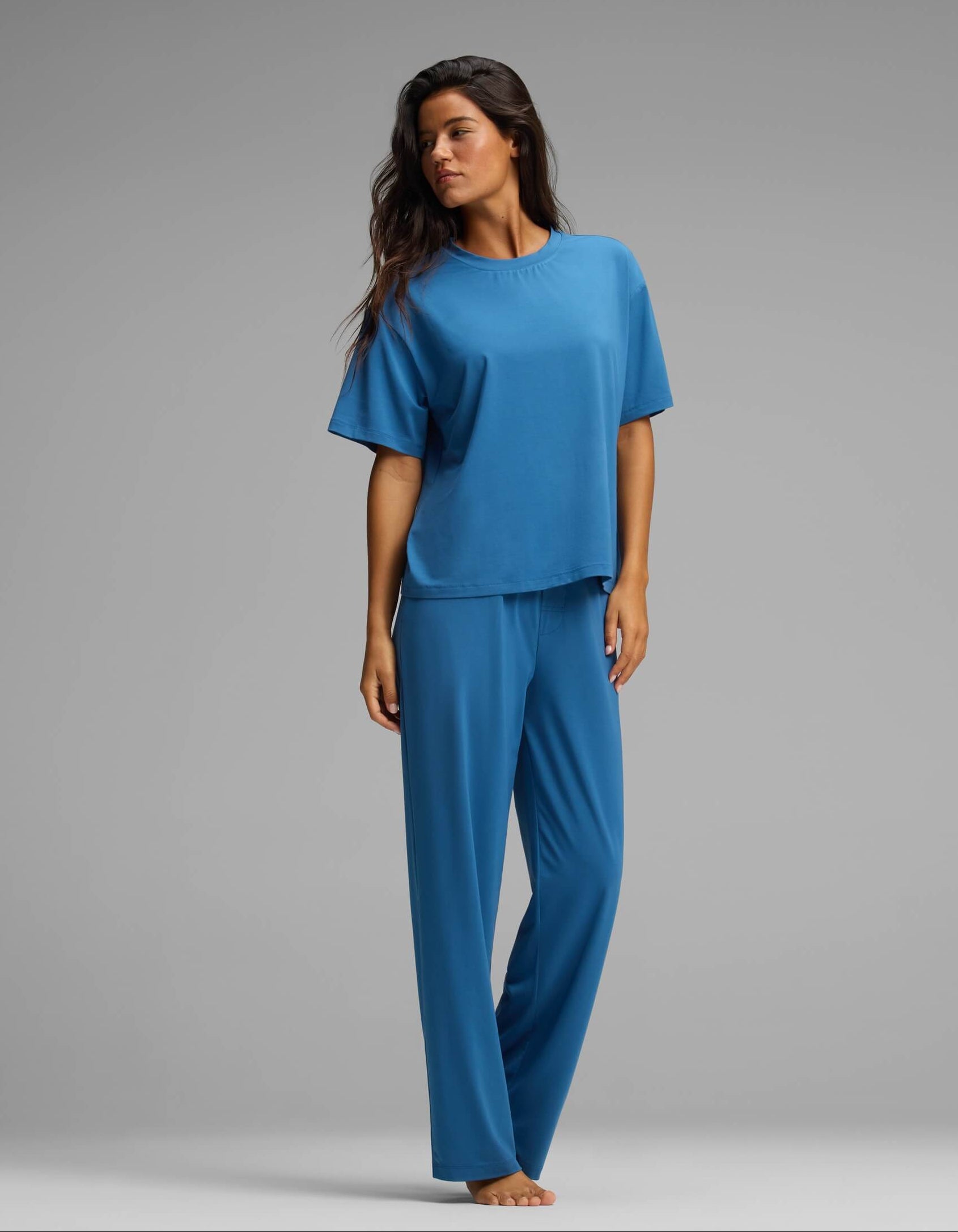 A woman stands barefoot against a gray background, wearing the Cozy Earth Women’s First Layer Pajama Short Sleeve Tee & Pant Set in a relaxed, loose-fitting style, looking to the side with an easygoing posture.
