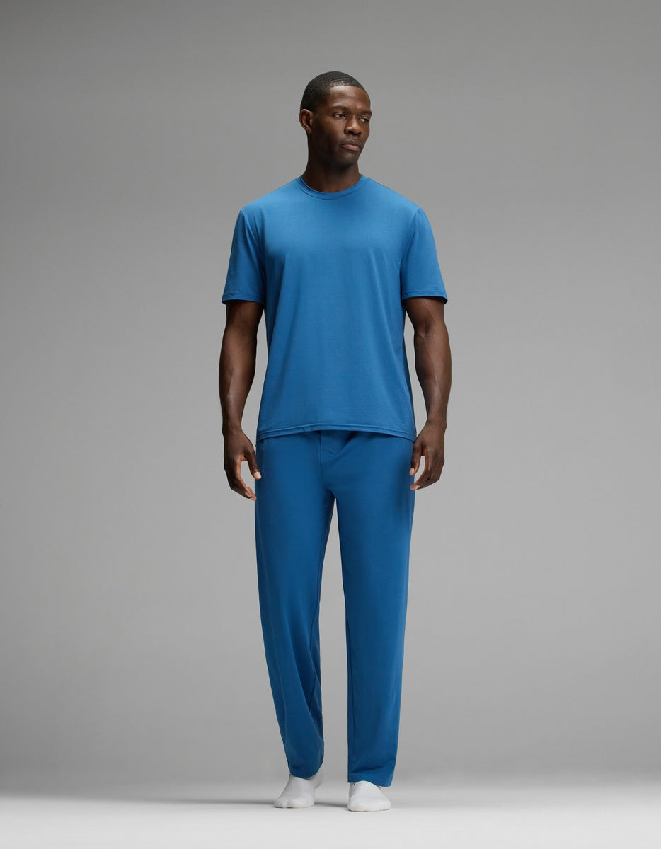 A man stands against a gray background wearing the Cozy Earth Men’s First Layer Pajama Short Sleeve Tee & Pant Set in blue with white socks, looking slightly to the side.